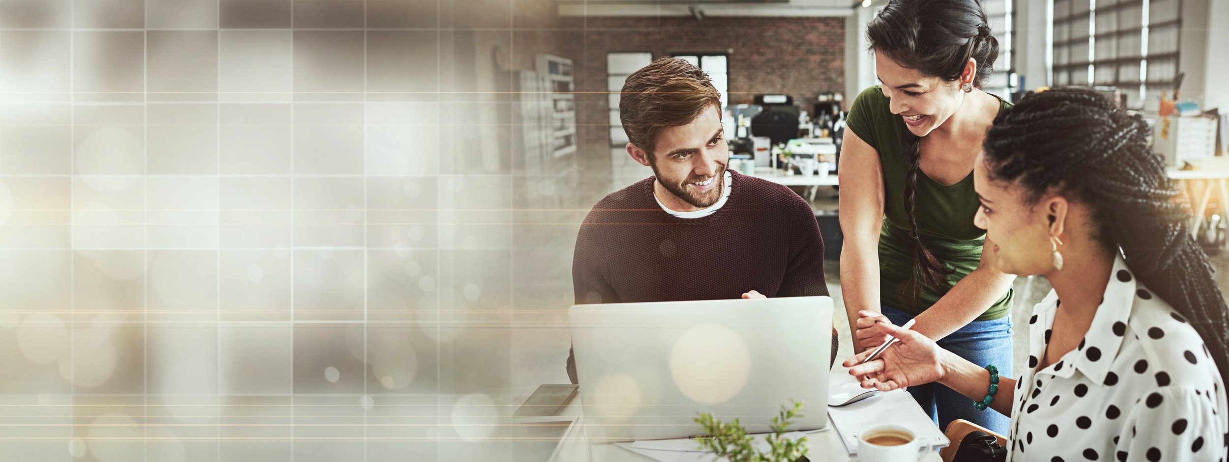 Three colleagues discussing over a laptop desk in an office with brick walls and large windows.