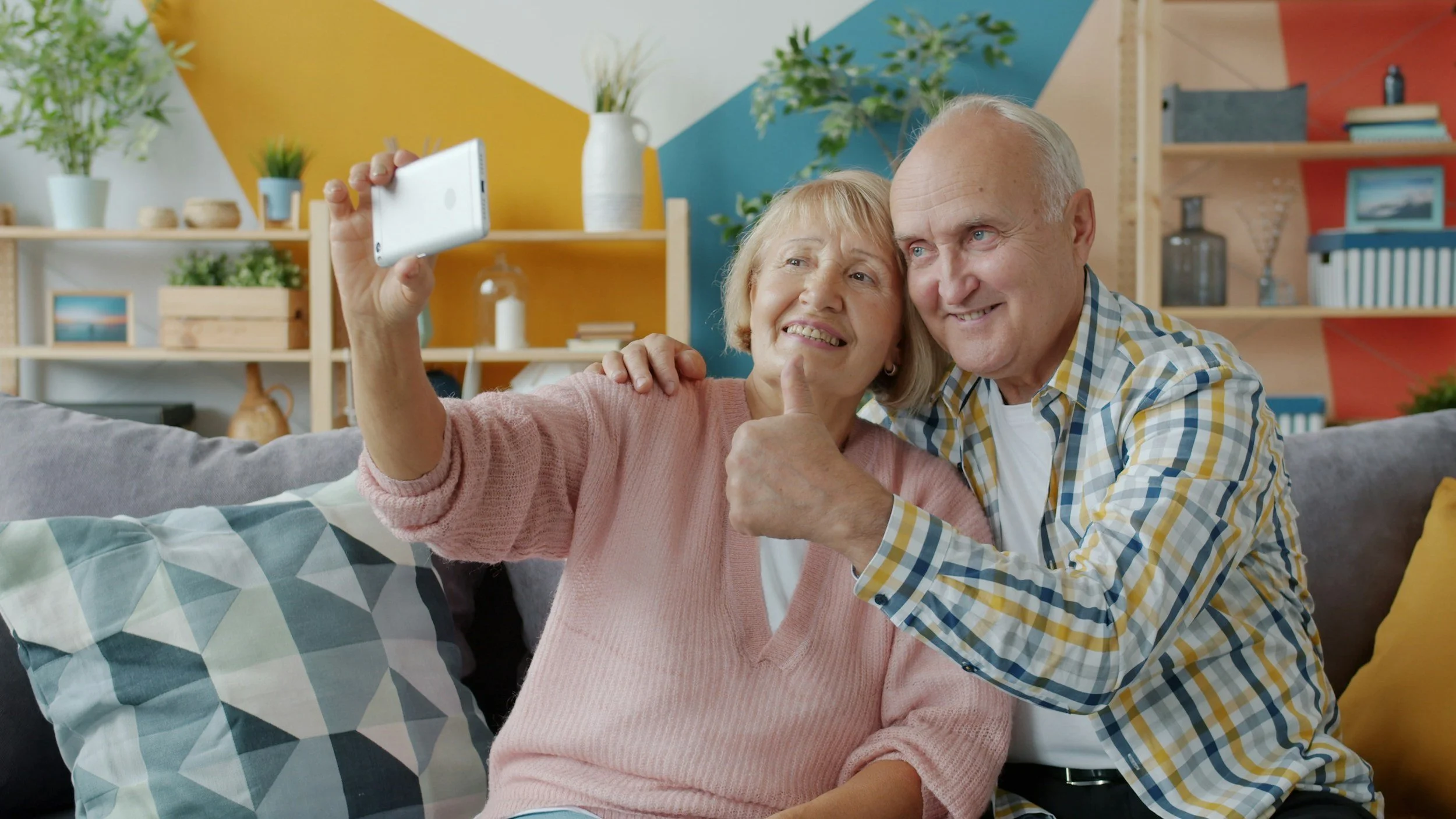 An elderly couple taking a selfie on a smartphone in a colorful, well-decorated living room.