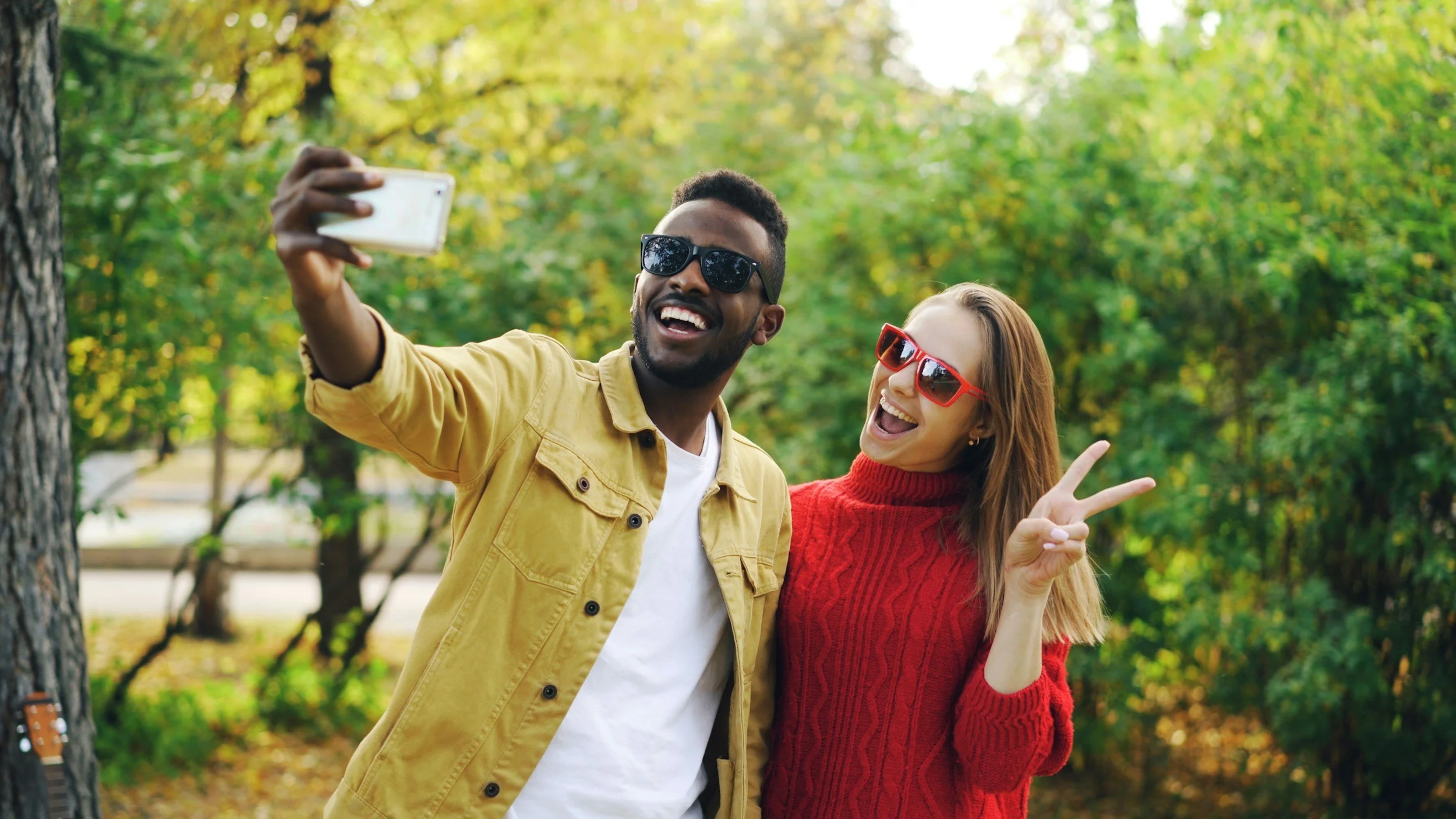 A man and woman taking a selfie outdoors with trees in the background, both wearing sunglasses and smiling. The woman is making a peace sign.