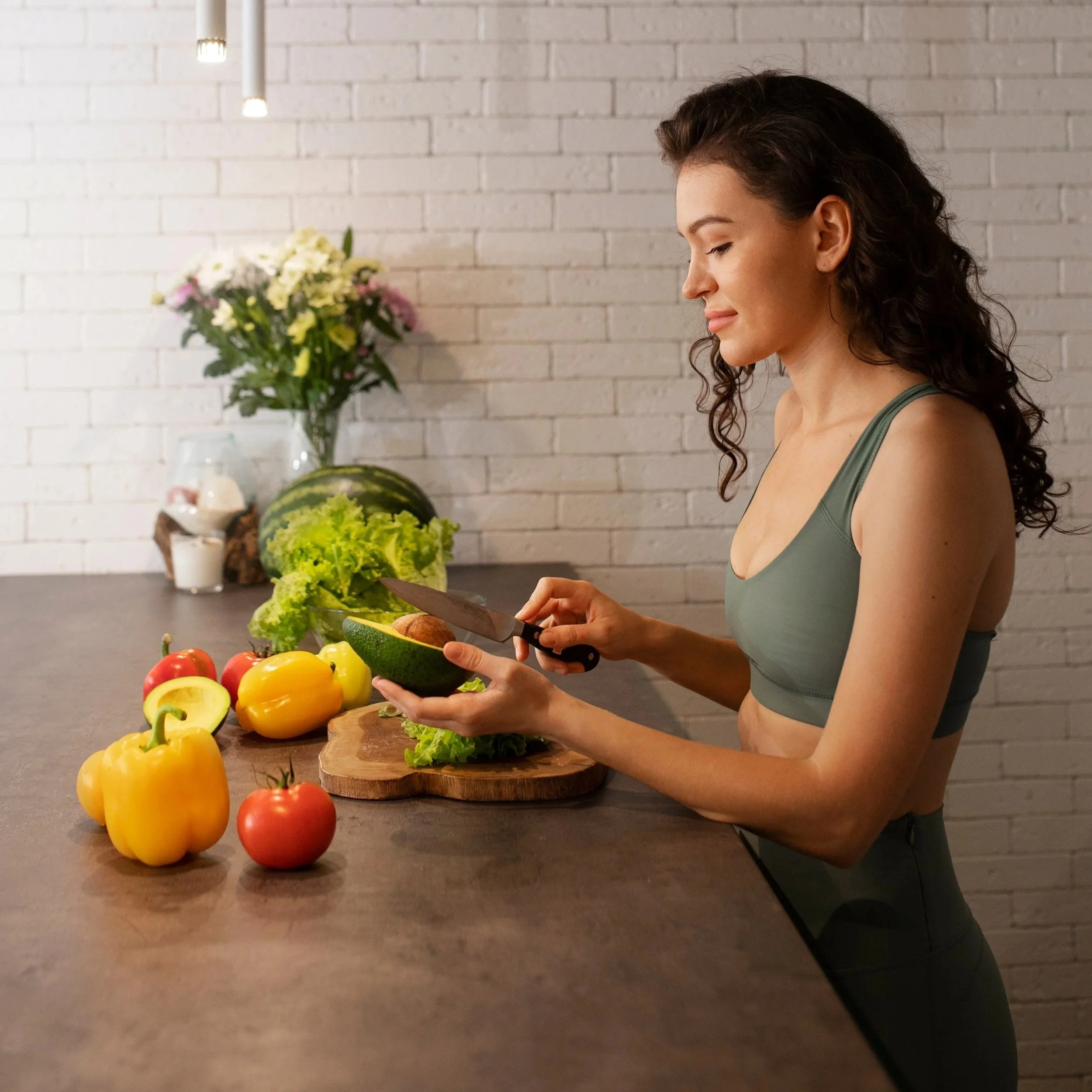 A woman preparing vegetables in a modern kitchen, with a variety of fresh peppers, an avocado, and leafy greens on the counter.