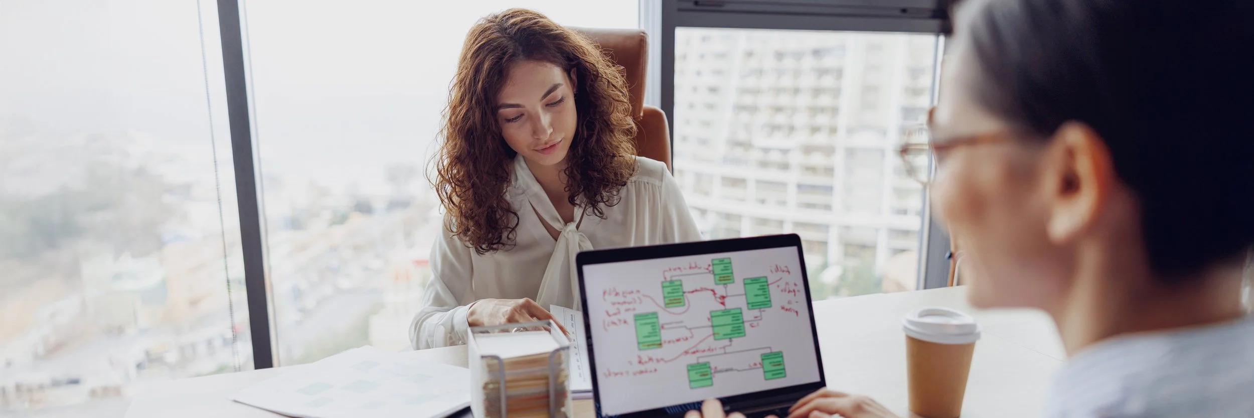 Two women in a modern office discuss documents and a laptop with a flowchart, with large windows showing a cityscape in the background.