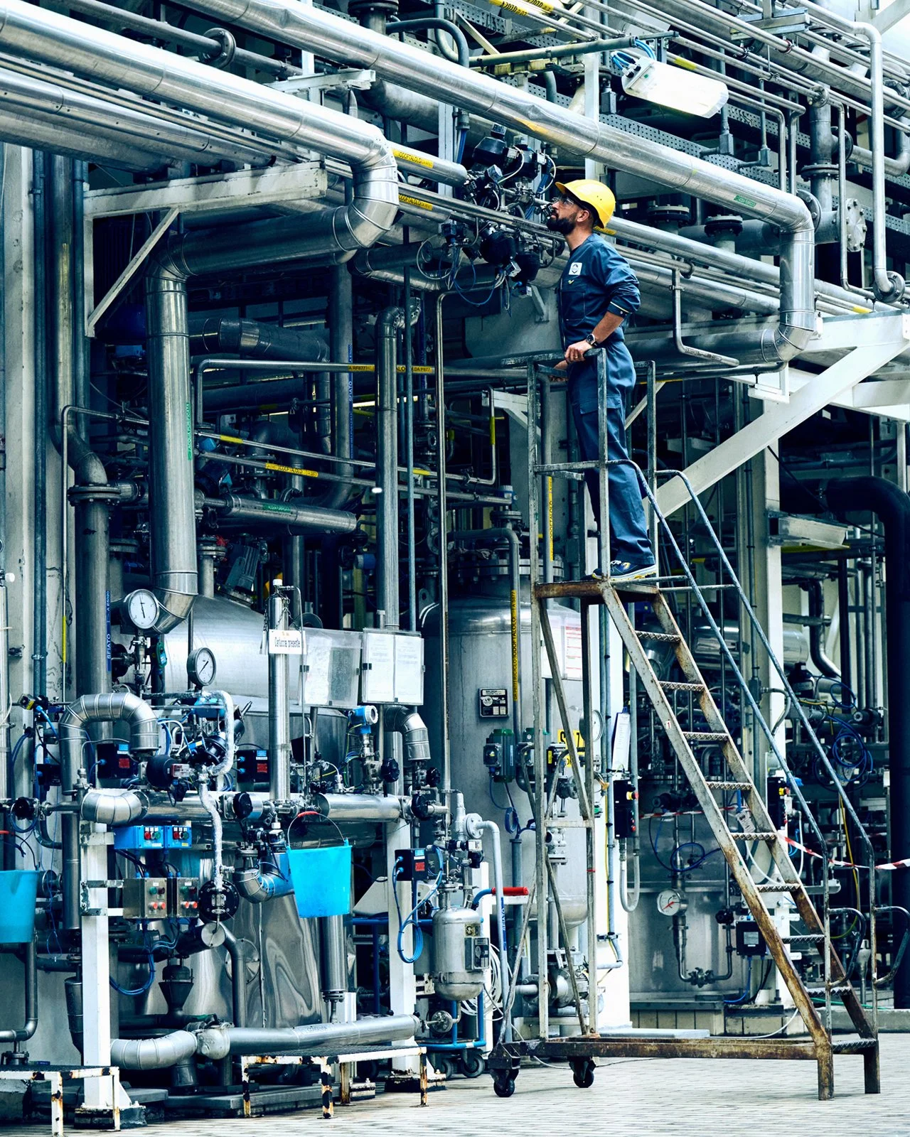 A worker in safety gear inspecting industrial pipelines and equipment, standing on a ladder in a factory setting.