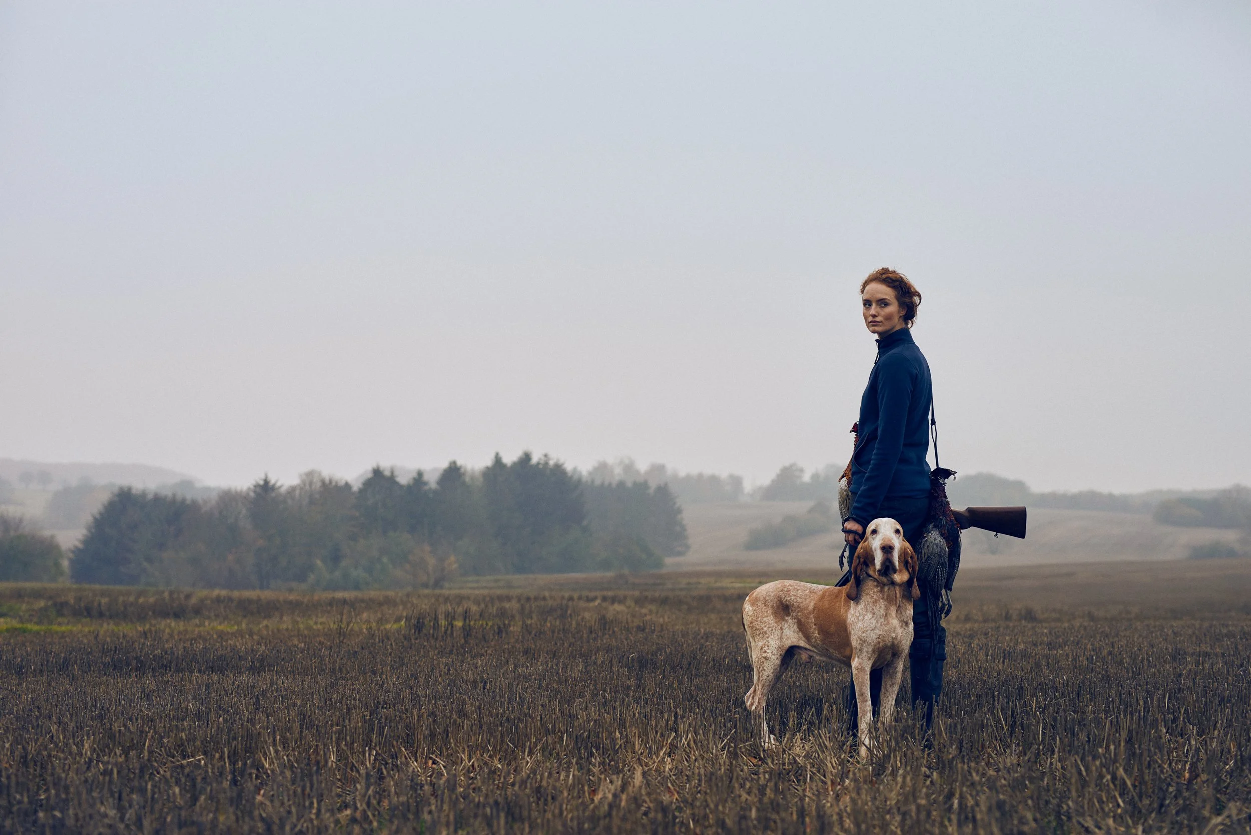 Woman standing in open field with hunting dog, wide landscape and overcast sky