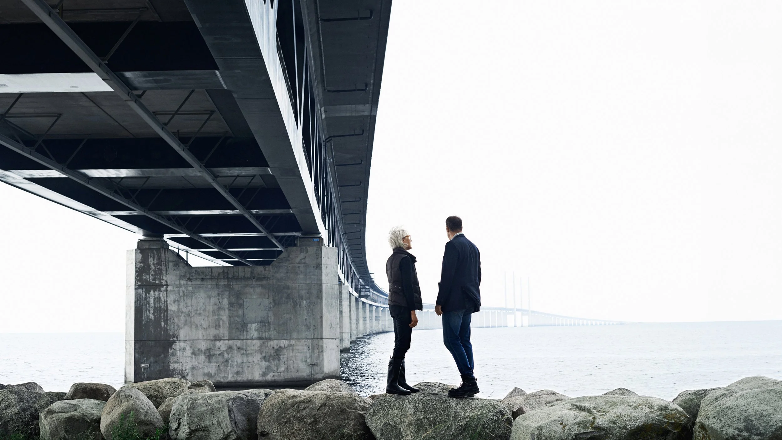 Two people in coats, a woman with short white hair and glasses and a man, stand under a large bridge by the water, facing each other.