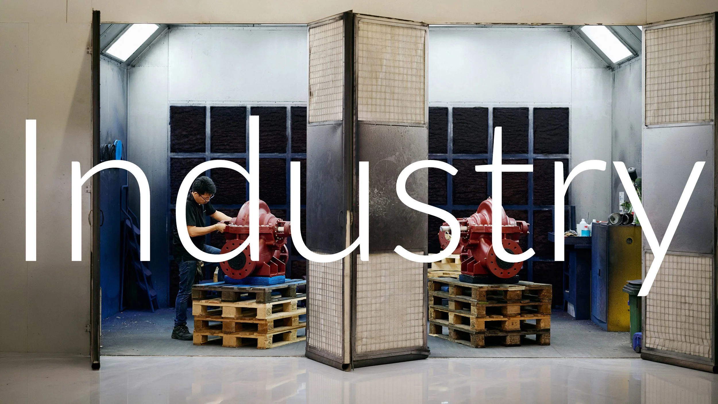 A person working on a red mechanical device inside a room with partition walls, with pallets and tools around.