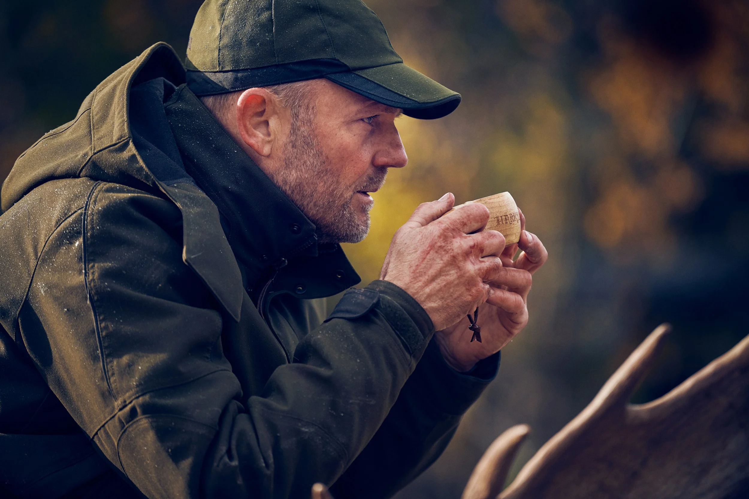 Hunter drinking from cup near fire, warming hands in outdoor setting