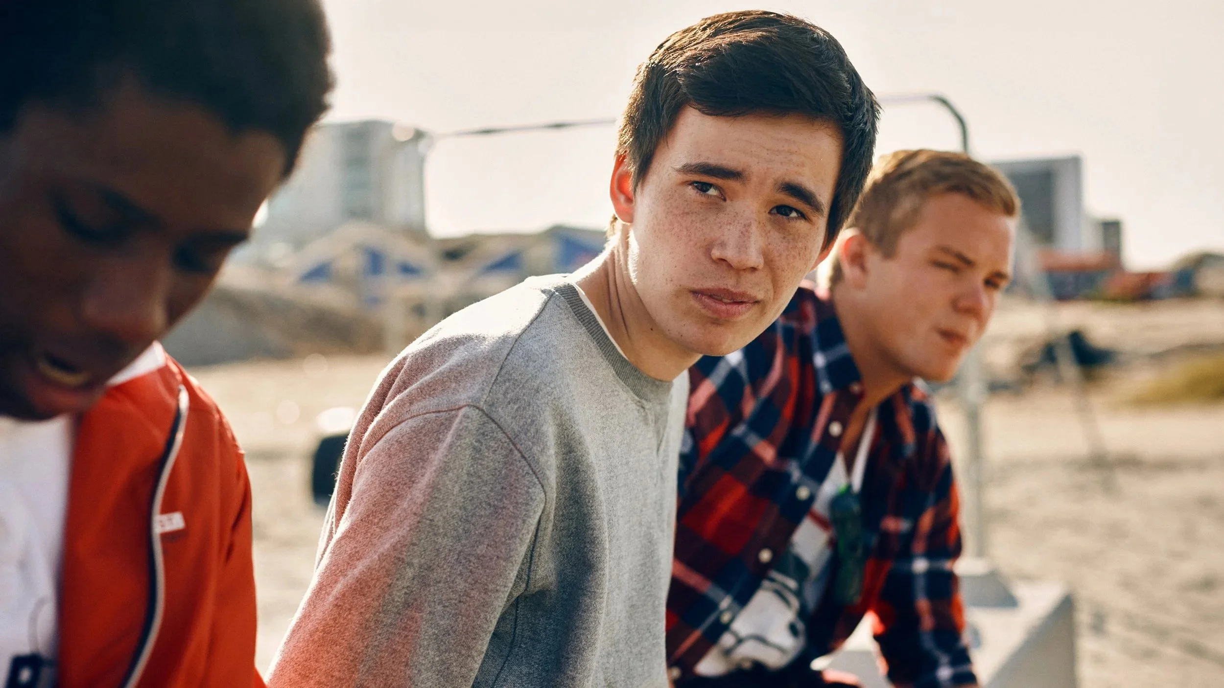 Three teenage boys sitting outdoors, one with a slightly confused or concerned expression, in a casual setting with buildings and boats in the background.