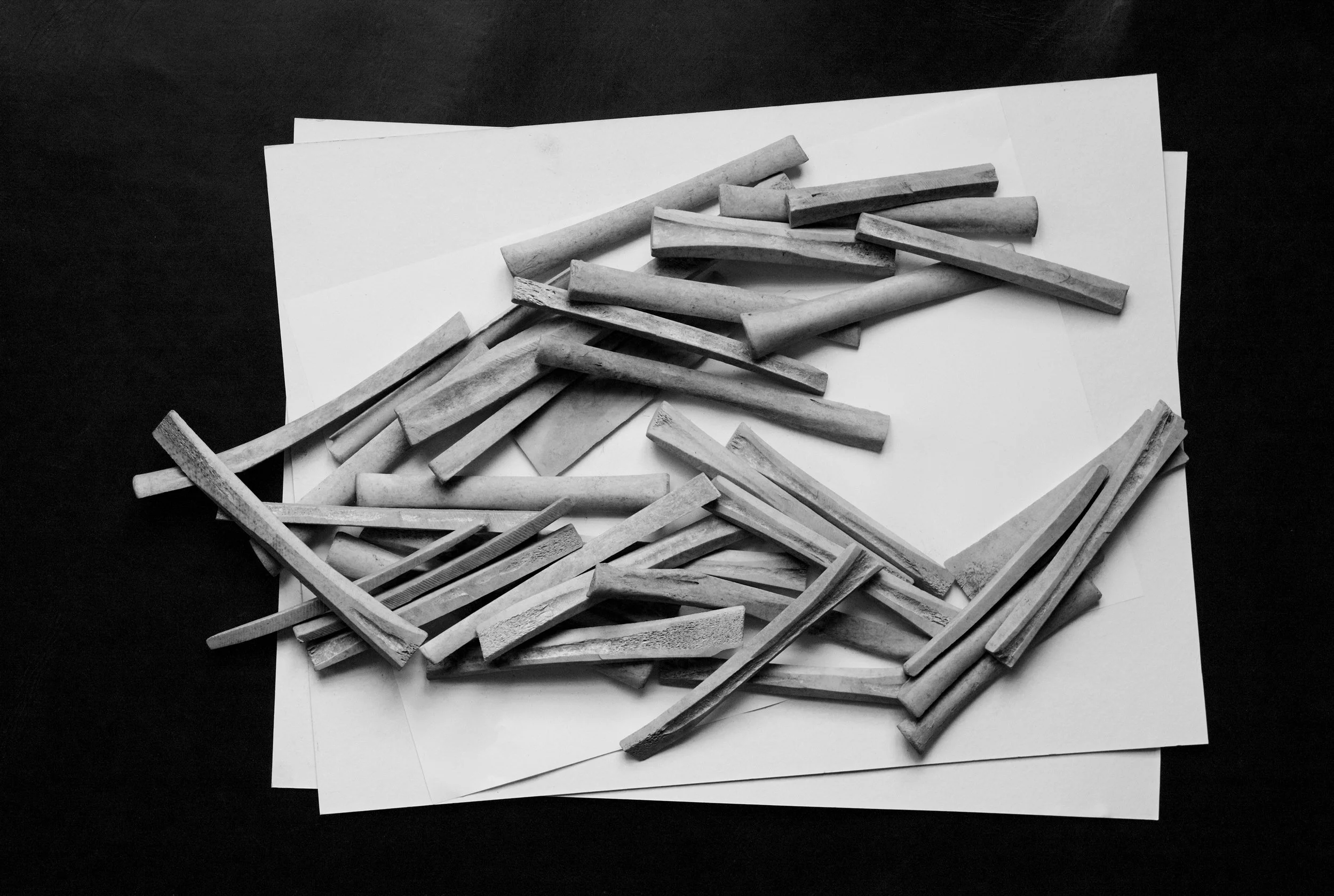Black and white photograph of scattered chalk sticks on sheets of white paper on a dark background.