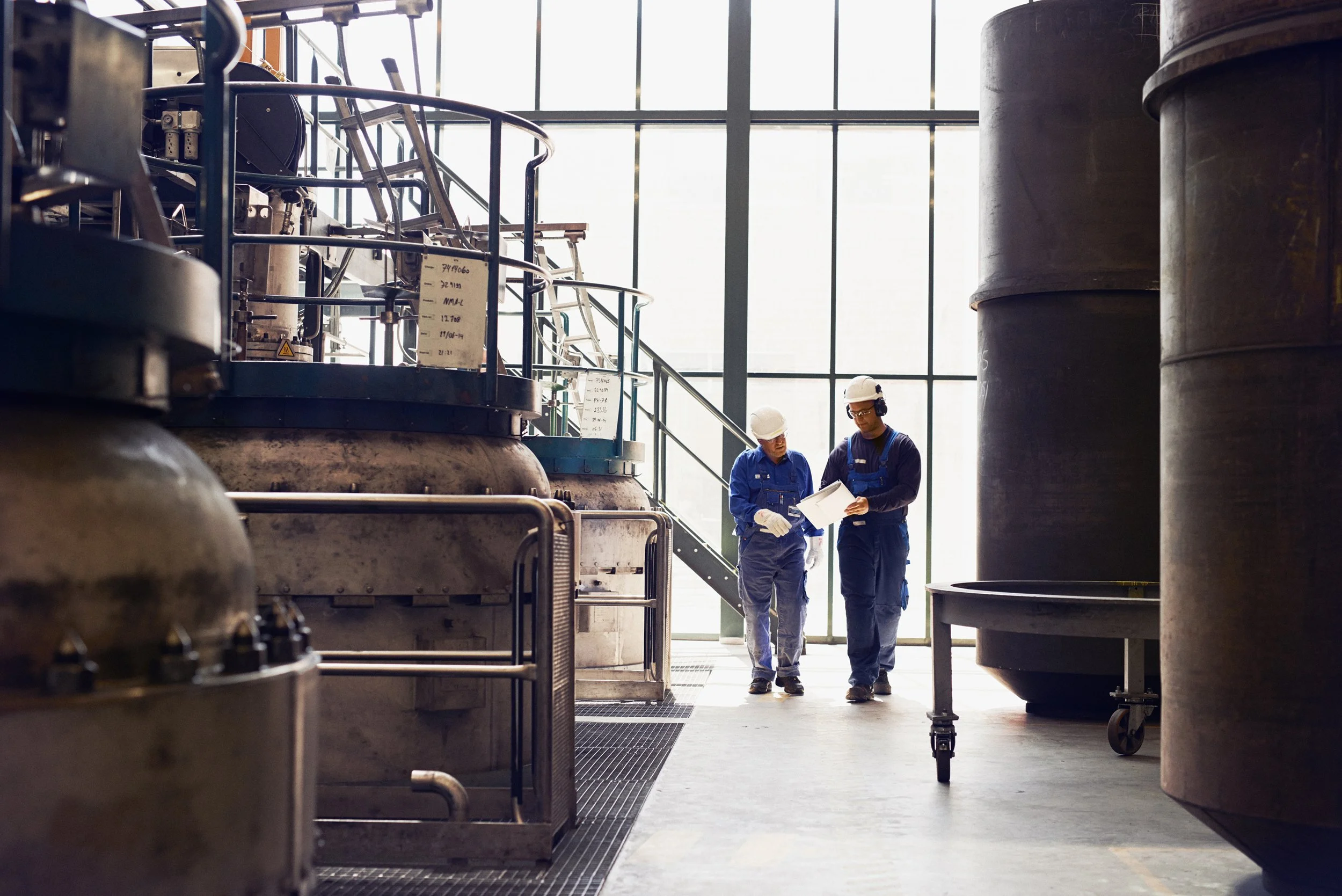 Two technicians reviewing documents while walking through industrial production facility between large processing units
