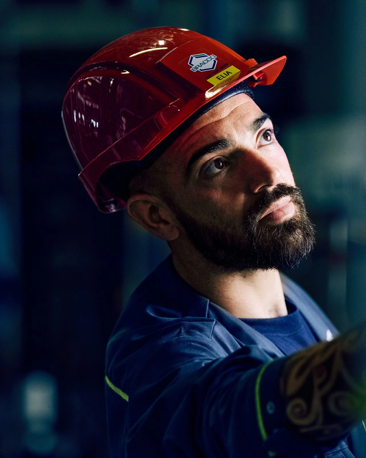 A male worker wearing a red safety helmet with a company sticker, looking upwards in a dimly lit industrial or construction setting.