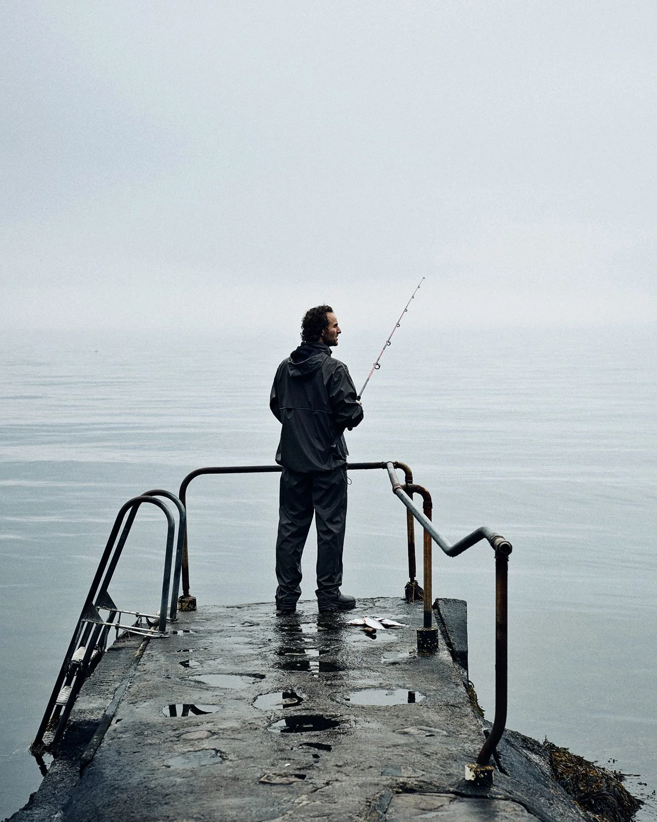 A man standing on a weathered pier in the water, holding a fishing rod, with a foggy, calm sea in the background.