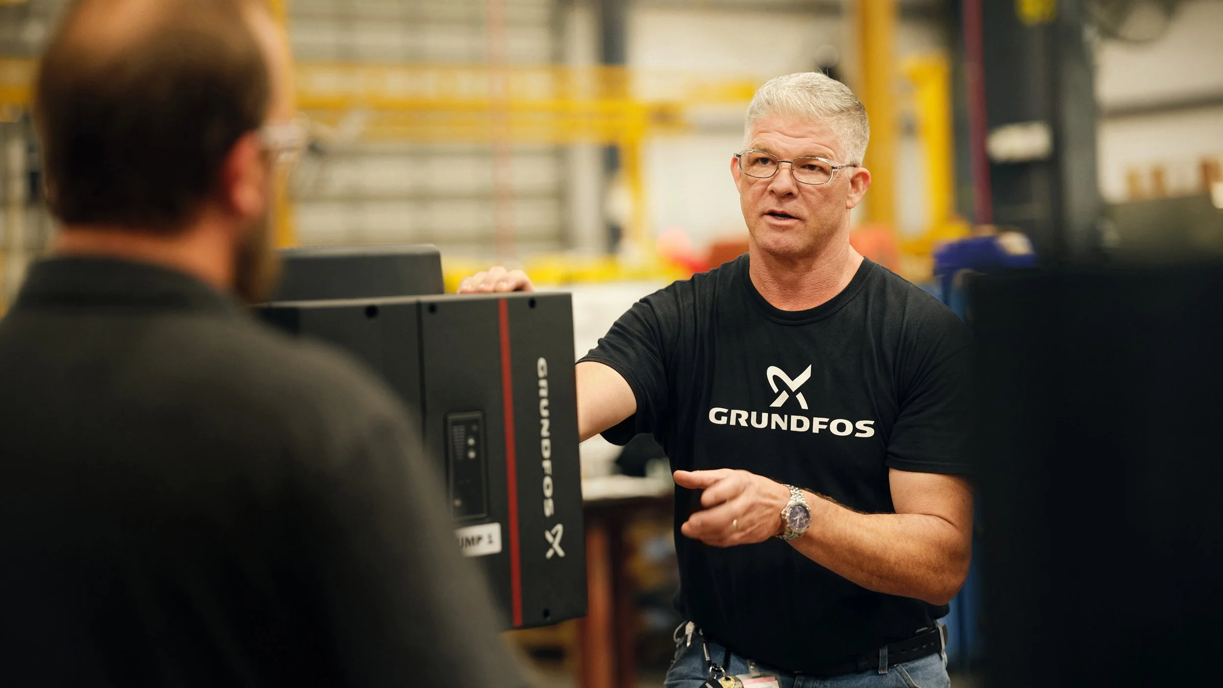 A man with gray hair, glasses, and a black Grundfos shirt is talking to another person in an industrial setting around machinery.