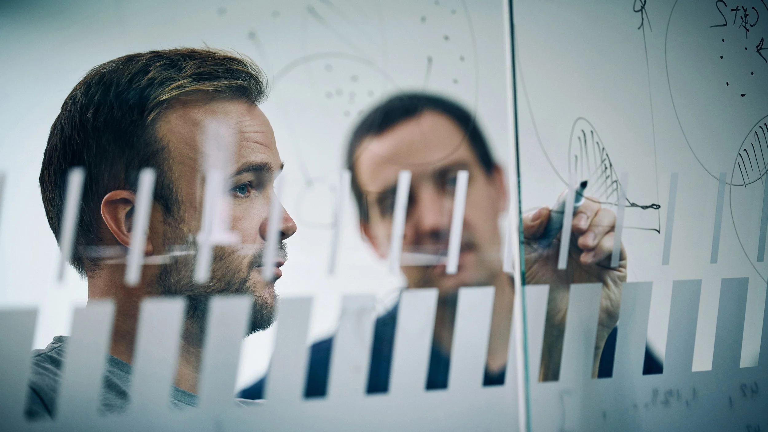 Two men are writing and drawing on a transparent glass or whiteboard with marker pens, with one man in focus and the other blurred.