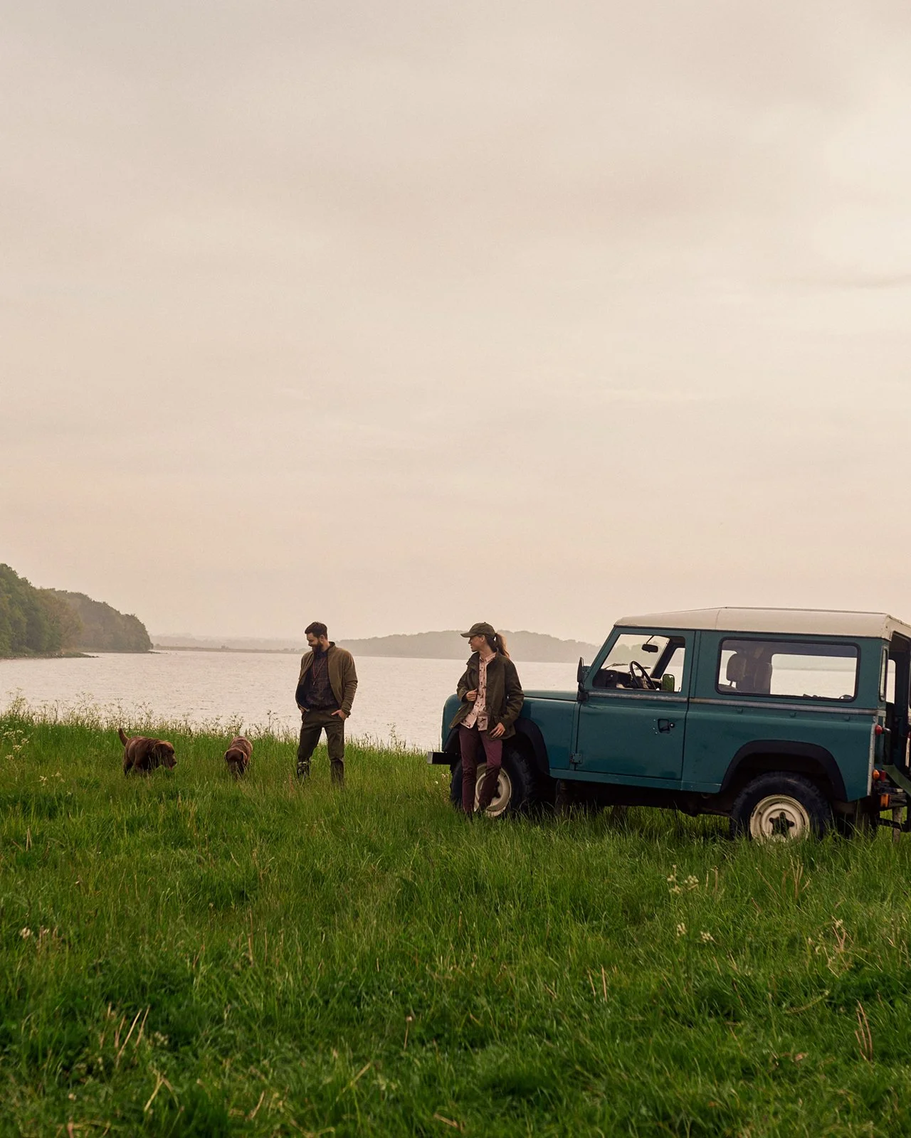 Two people standing on a grassy area near water, with a blue off-road vehicle and two dogs, under an overcast sky.