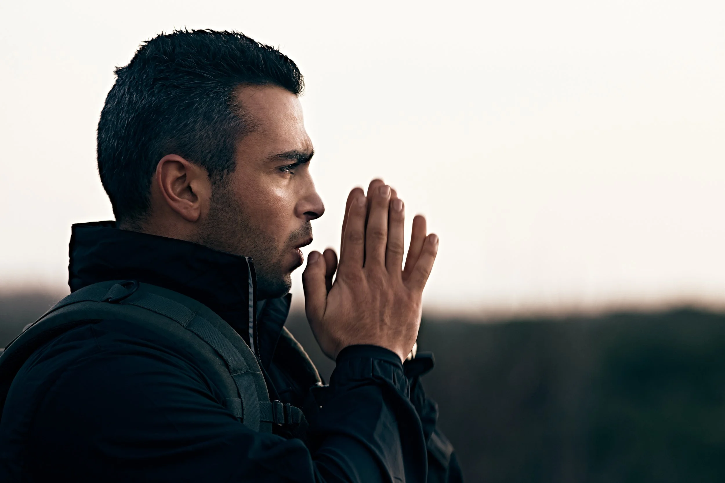 Man standing outdoors with hands together near face wearing jacket and backpack