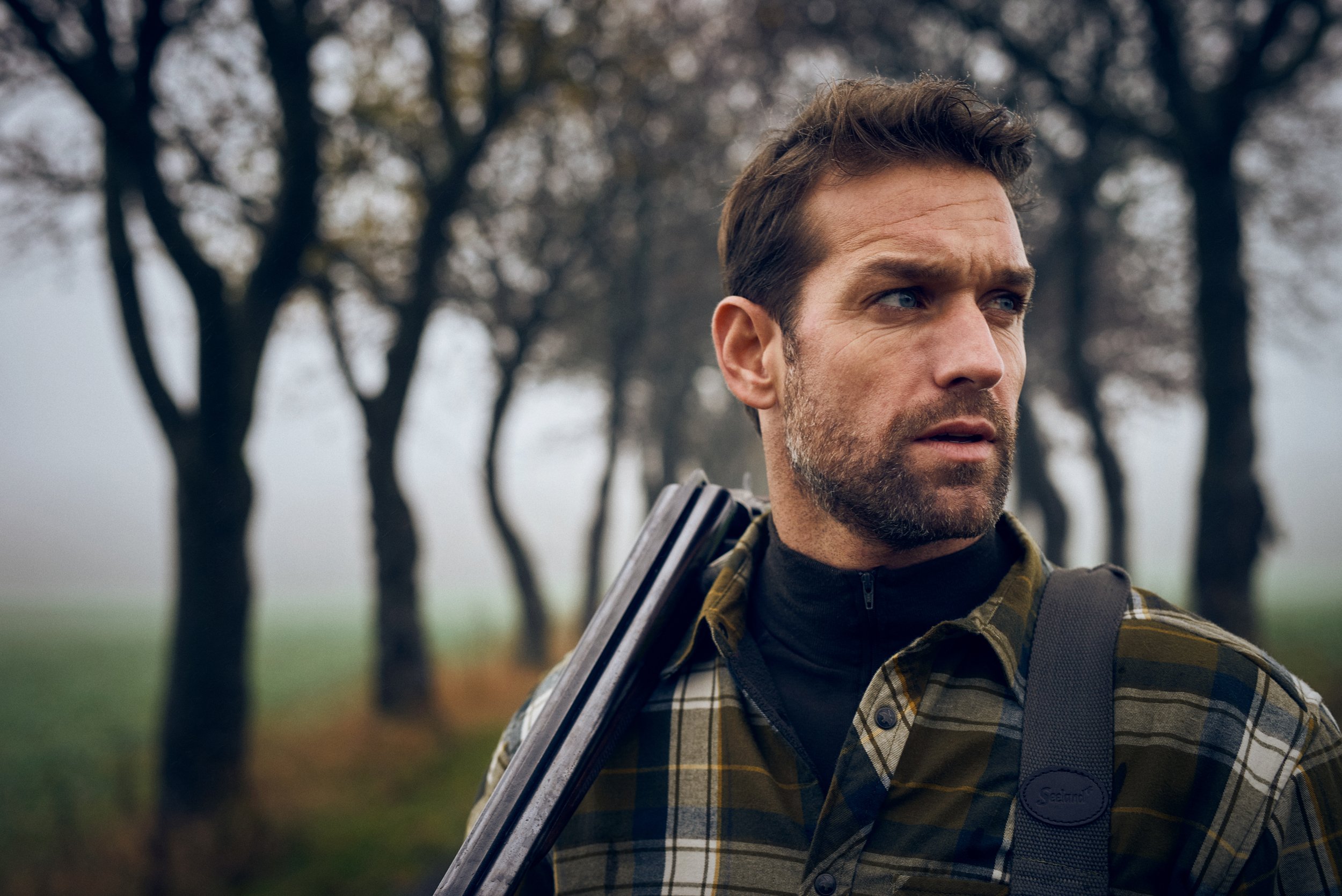 Portrait of man carrying shotgun over shoulder, focused expression along tree-lined path