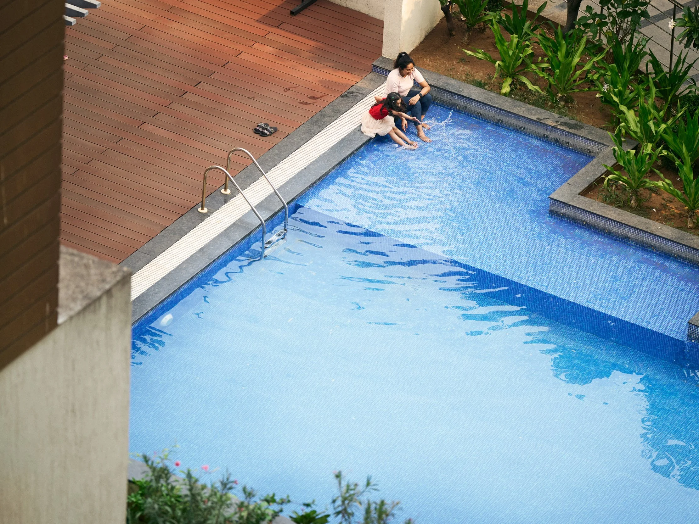 Two people sitting at edge of outdoor swimming pool in residential courtyard, viewed from above
