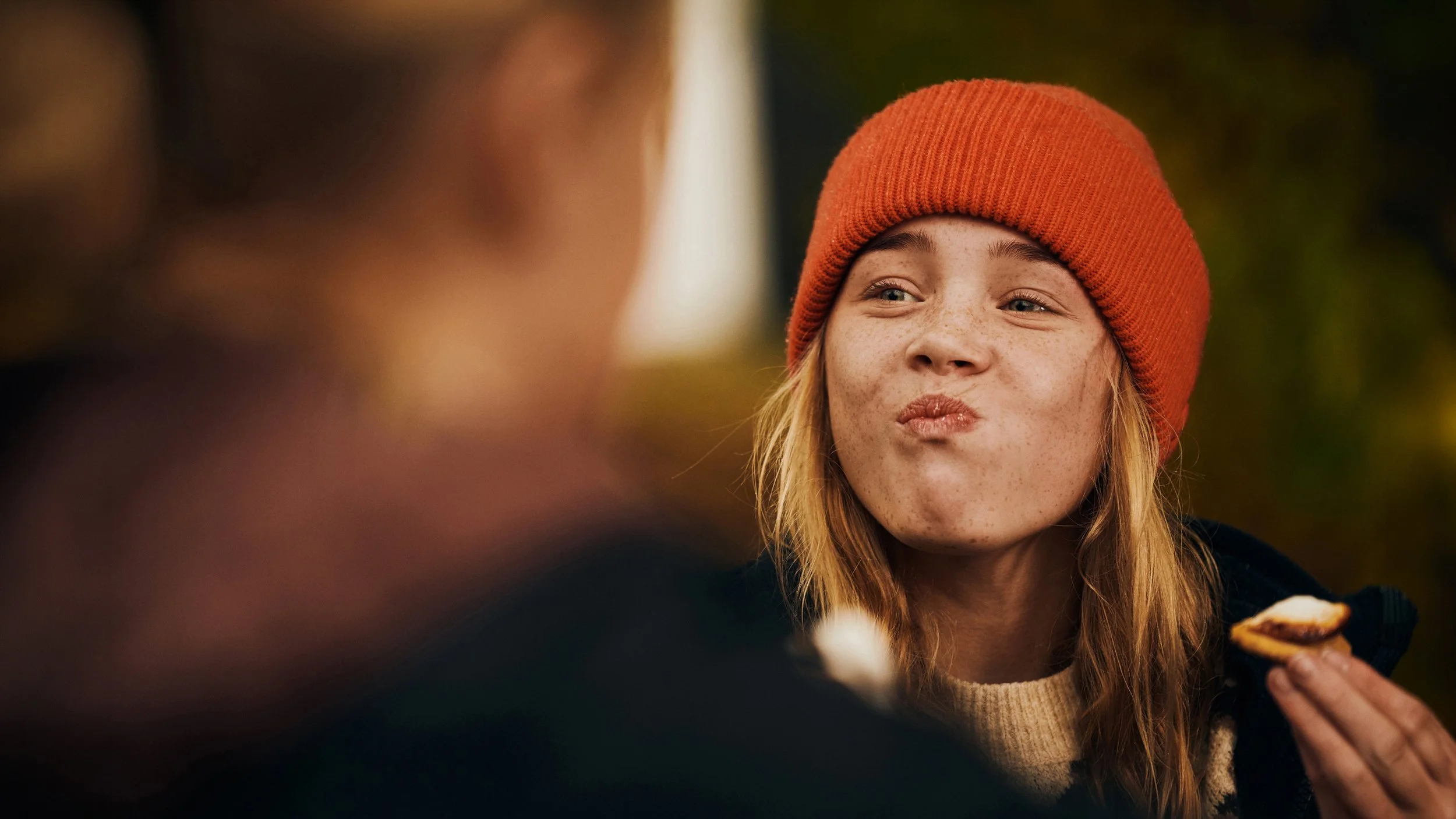 A girl with freckles wearing a red knit beanie and beige sweater, making a playful duck face while eating outdoors.