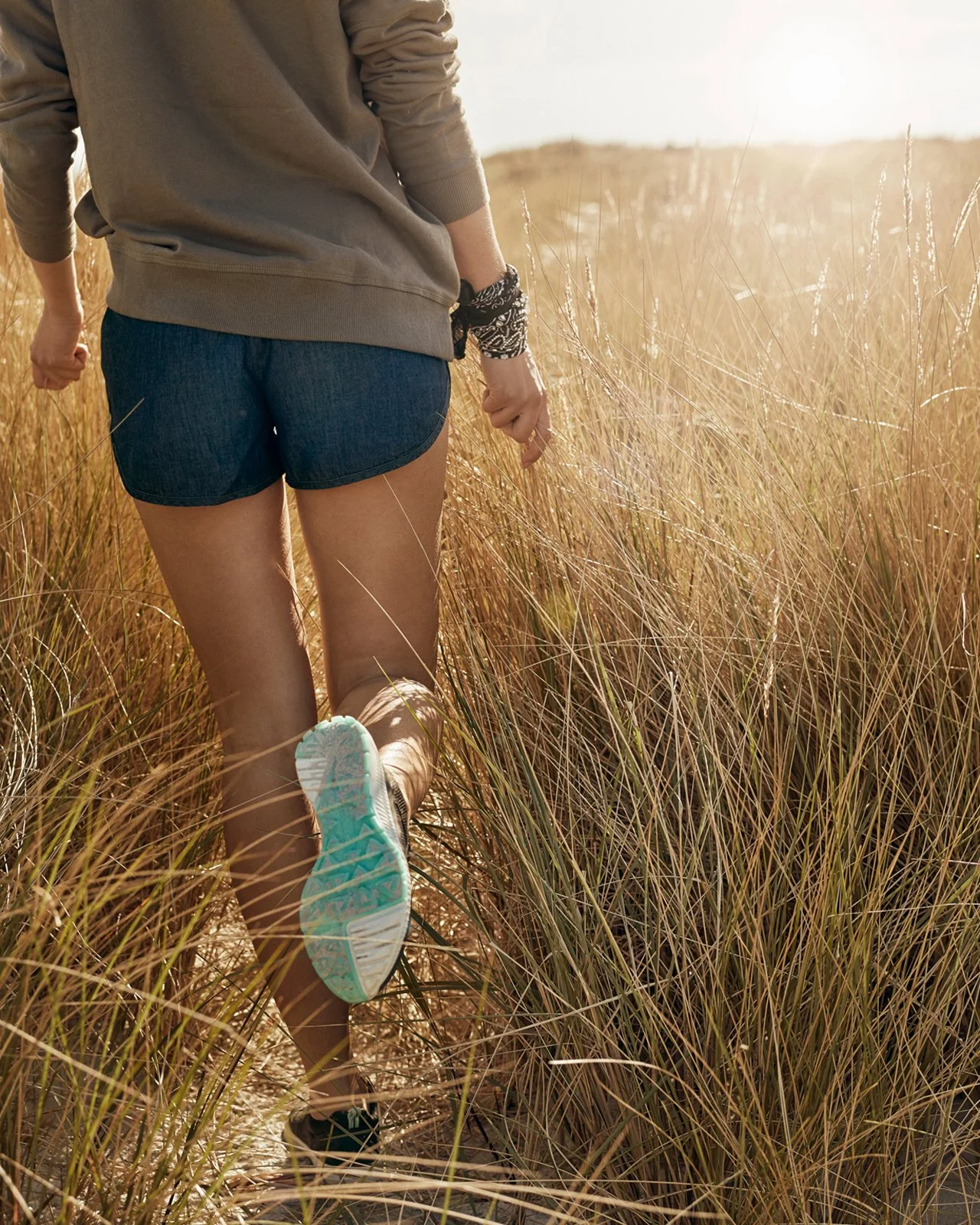 Person running through tall, dry grass at sunset, wearing a hoodie, shorts, and sneakers, with their back to the camera.