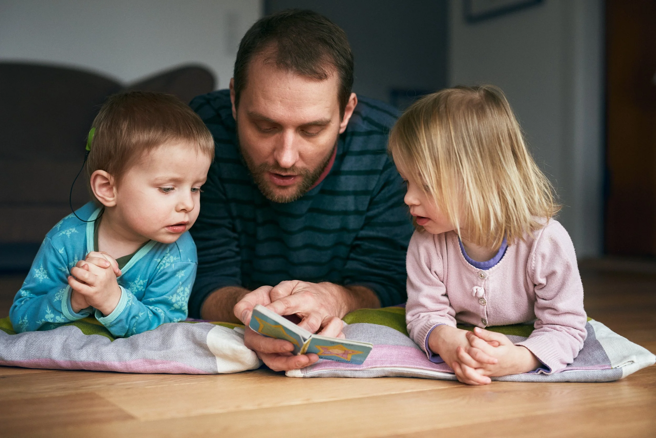 Father and two children with hearing aids reading together on floor, shared moment at home