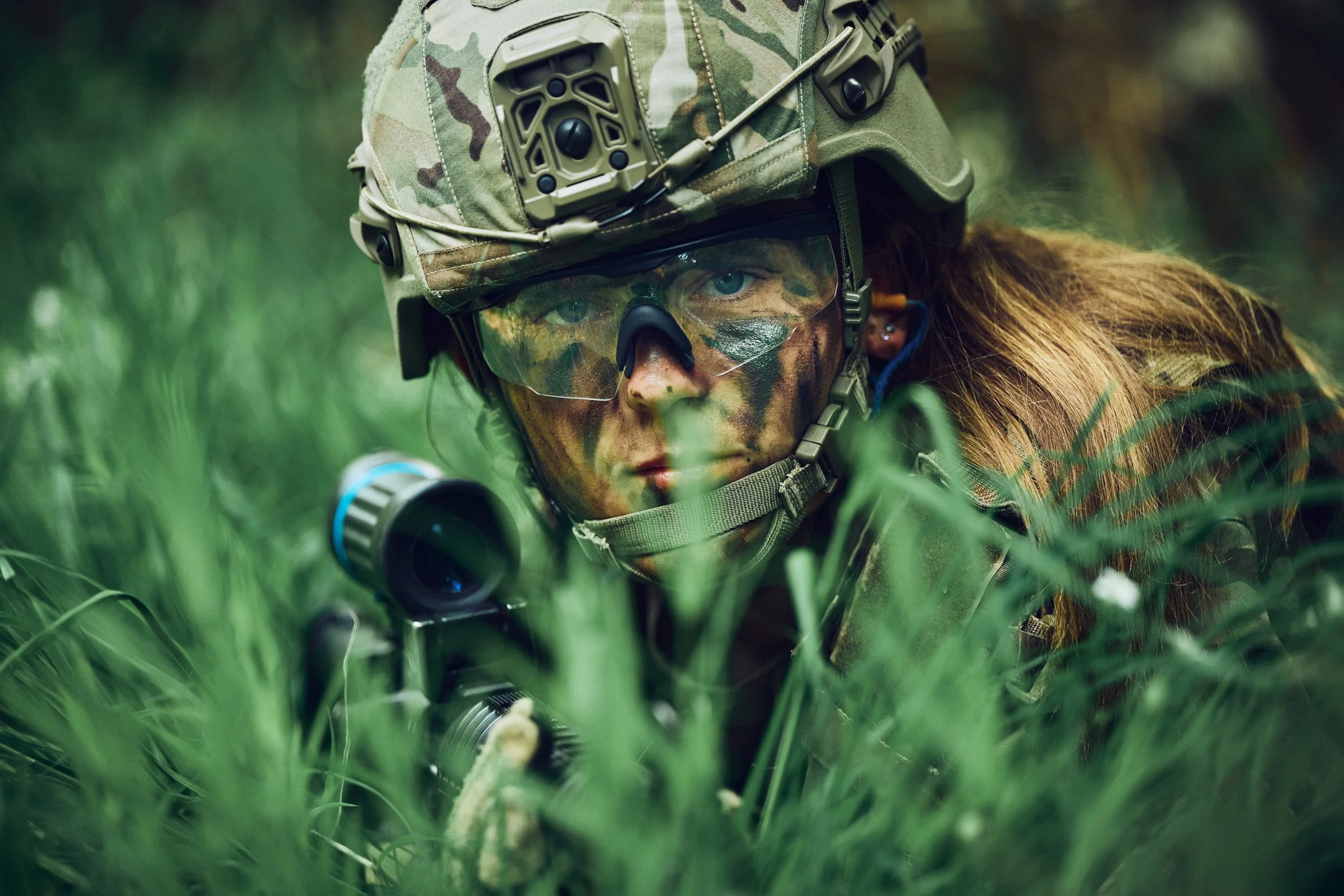 Camouflaged soldier lying in tall grass aiming rifle, face paint and tactical gear
