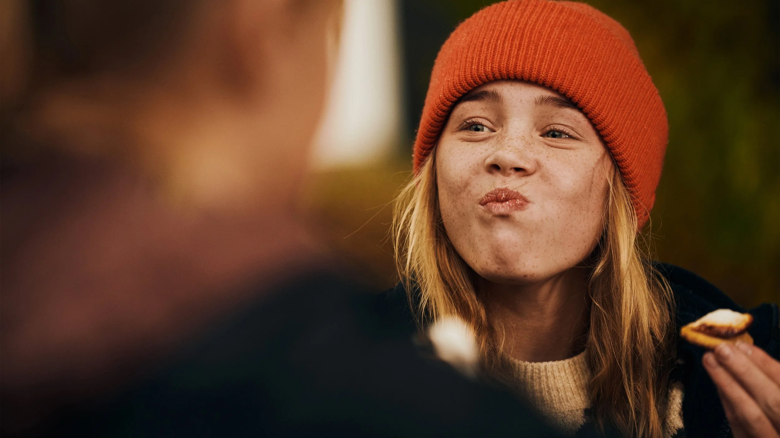 A young woman with freckles wearing an orange beanie is making a playful face while eating a cookie or pastry outdoors.