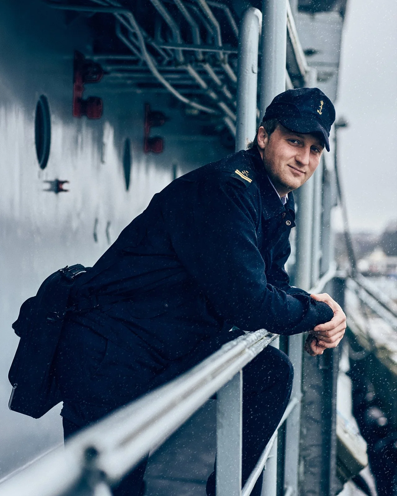A man in a navy uniform and matching cap is leaning on a metal railing outdoors, with a cloudy, rainy background and industrial elements behind him.