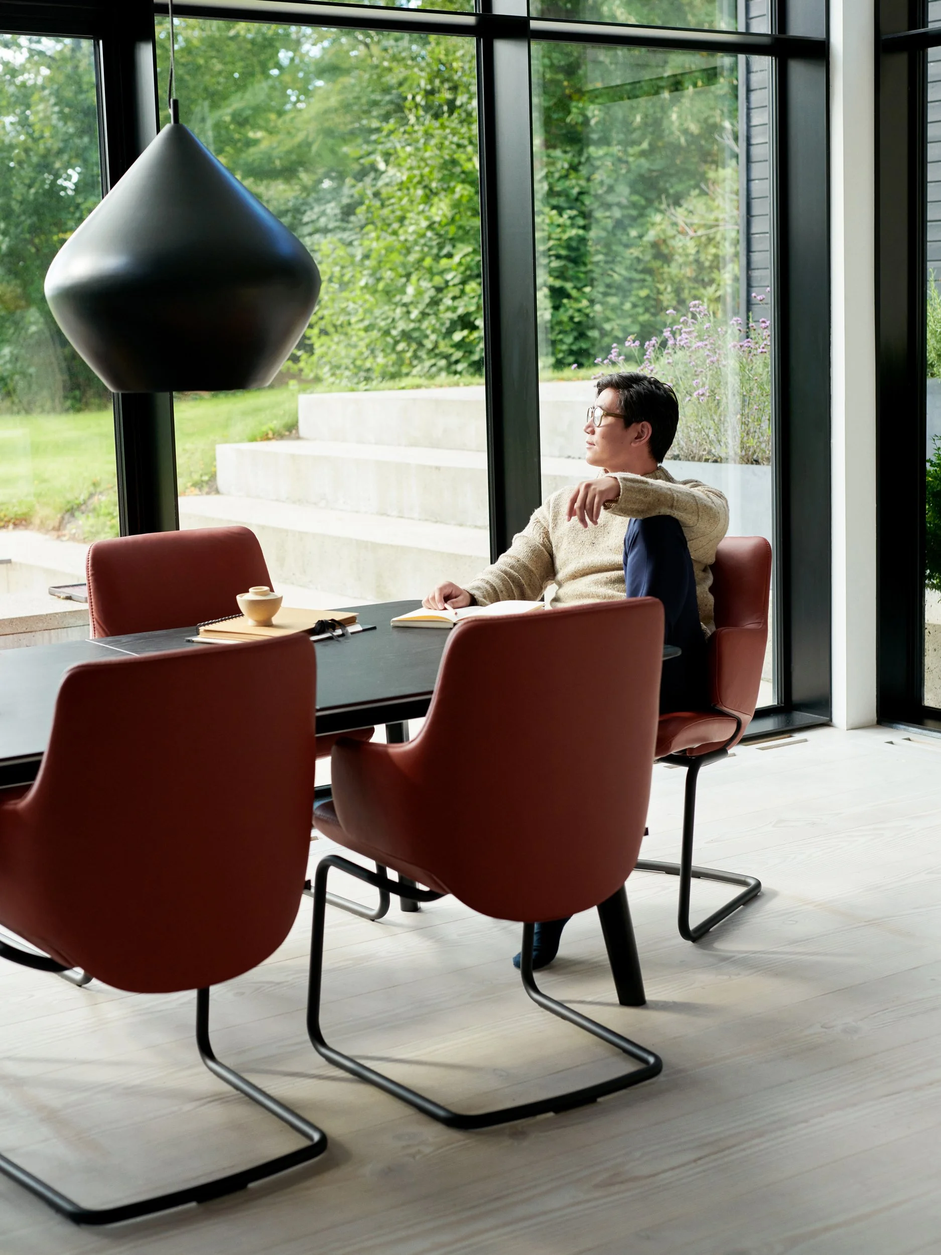 Man sitting at table by large window, looking out into garden, reflective moment