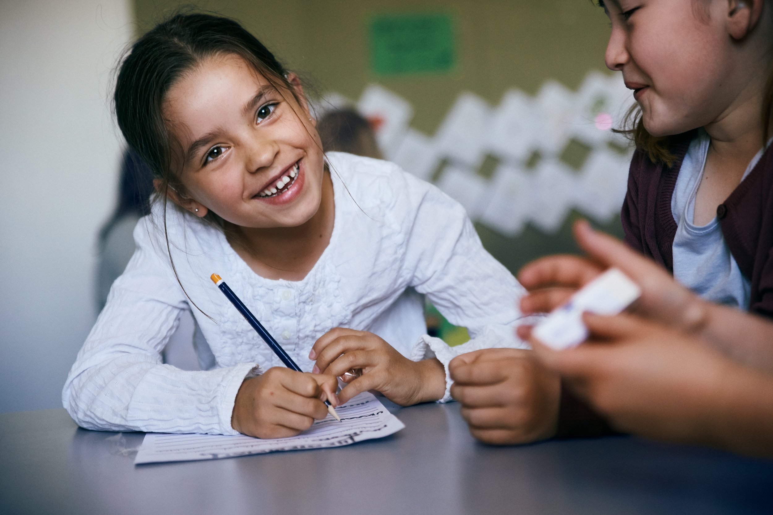 Child wearing hearing aids writing at table with others, social moment in classroom
