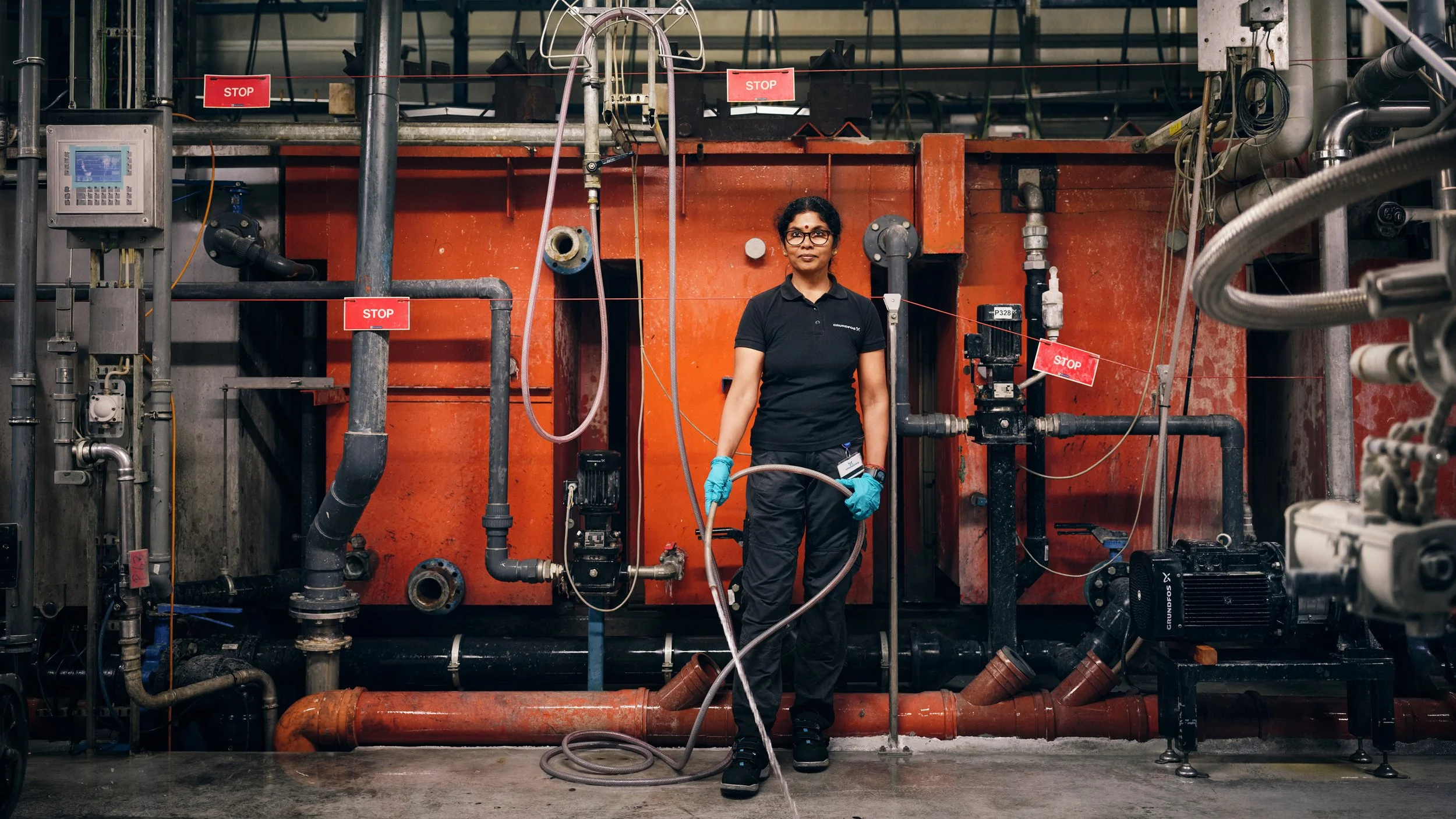 A woman wearing safety goggles, gloves, and a black shirt stands among industrial pipes and machinery, holding a hose in an industrial setting.
