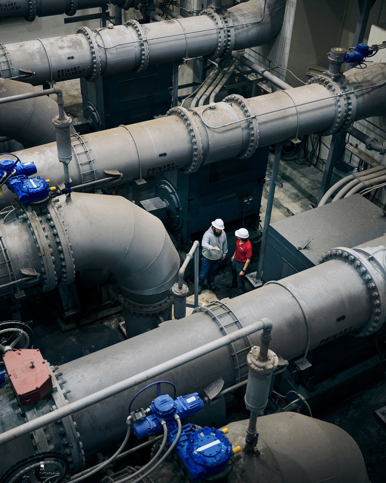 Two workers wearing white safety helmets and safety glasses are standing and talking among large industrial pipelines in an industrial facility.
