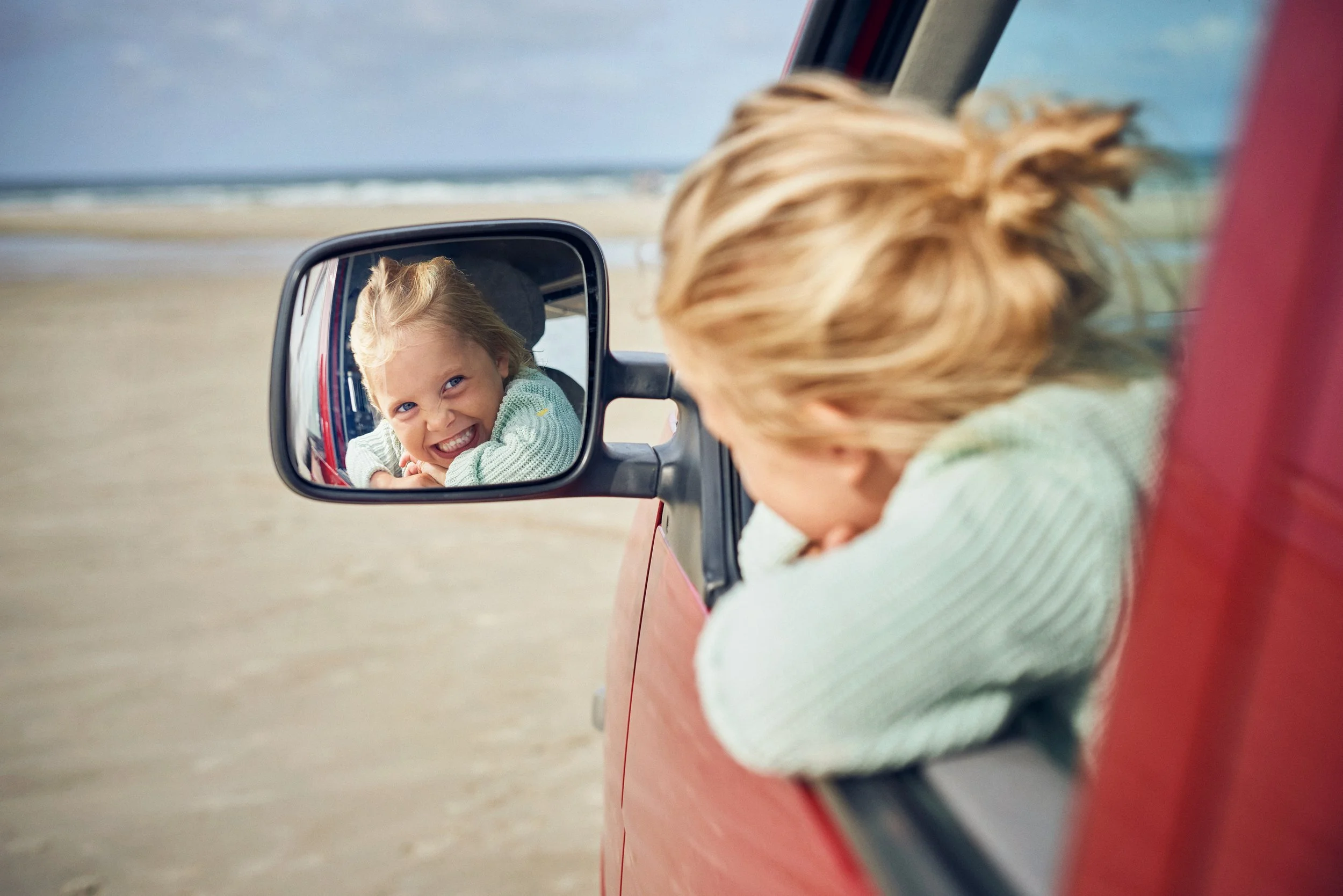 Child leaning out of car window looking into side mirror, playful travel moment