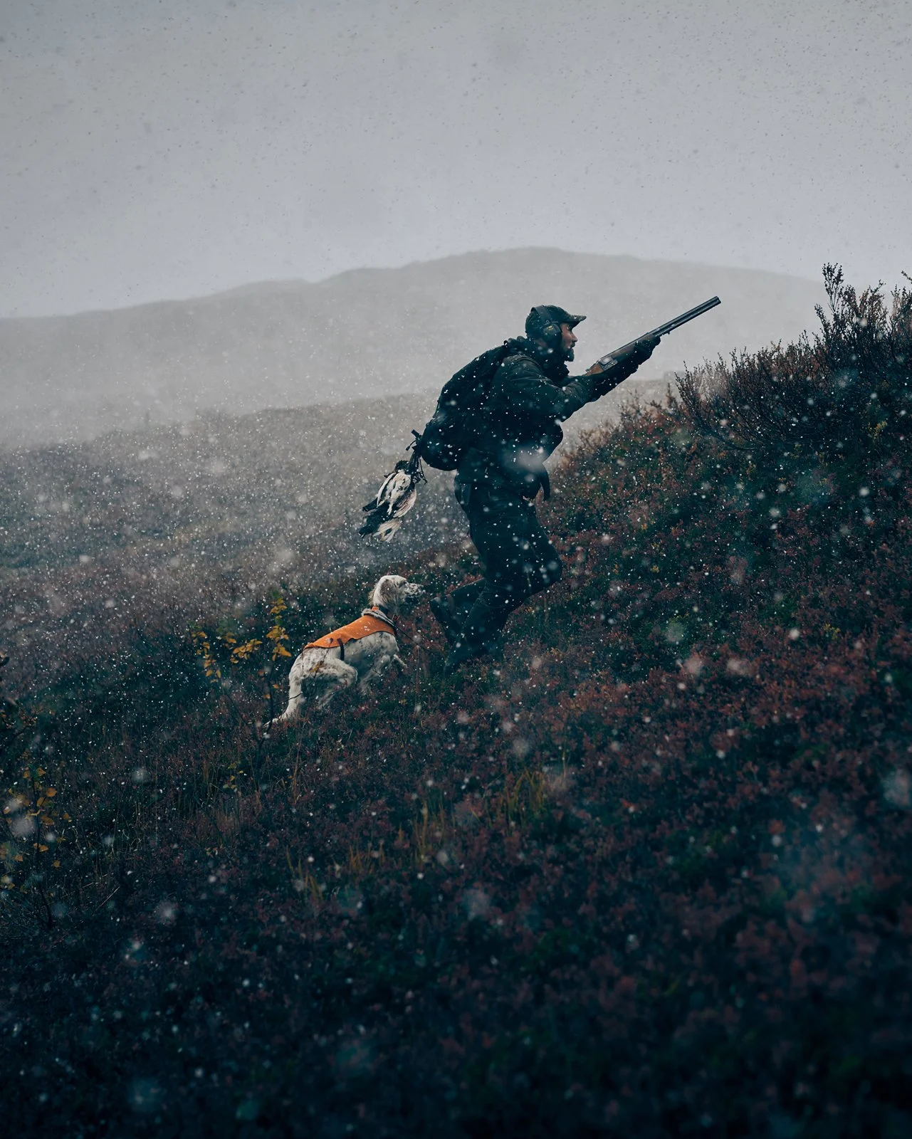 A man and his dog hiking through a snowy, mountainous terrain. The man is wearing outdoor gear and carrying a rifle, with the dog wearing an orange vest following closely behind.