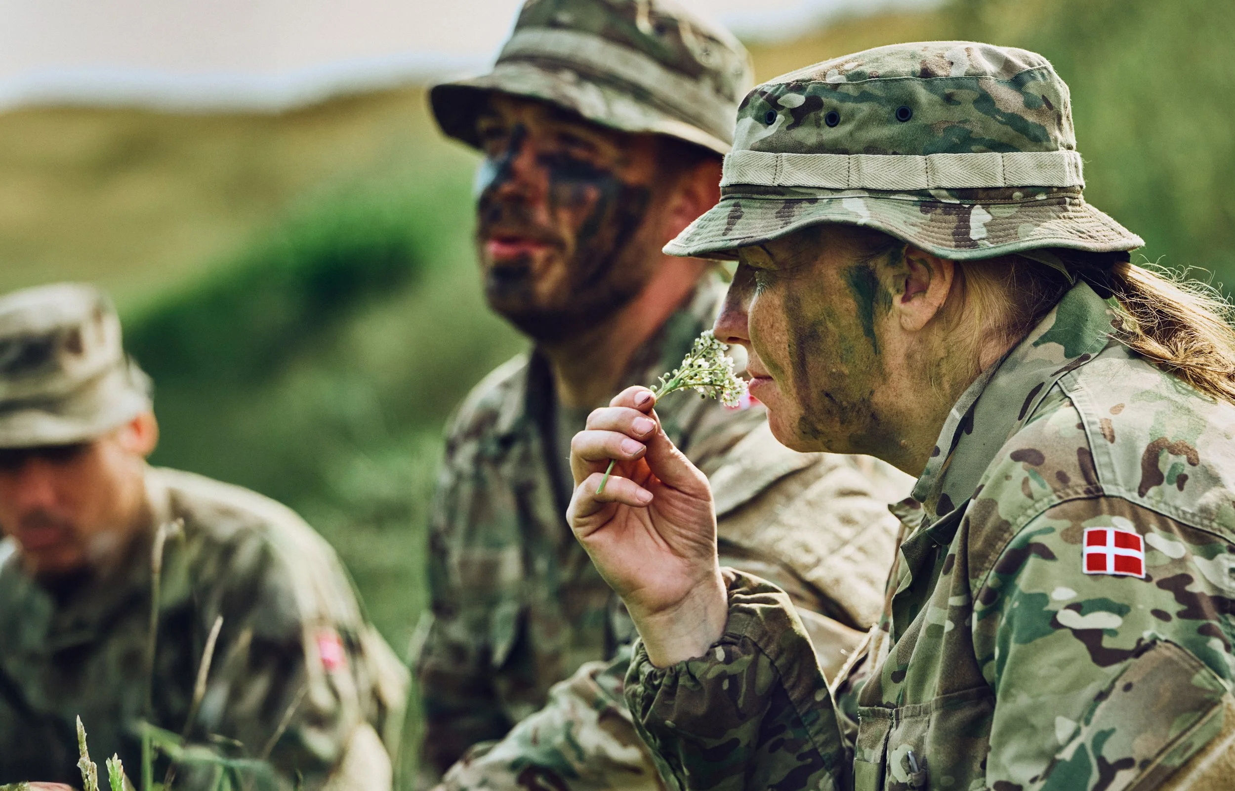 Soldiers with face paint resting in grass during exercise, one smelling plant in foreground