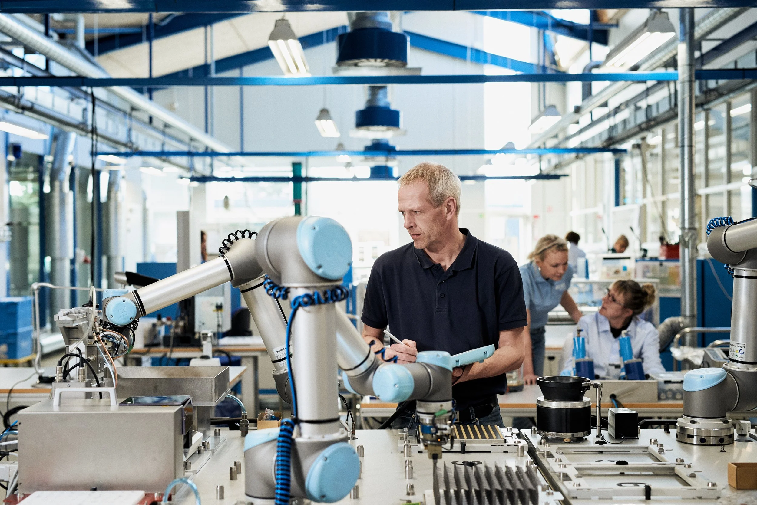 Technician operating collaborative robot arm in production environment while colleagues work in background 