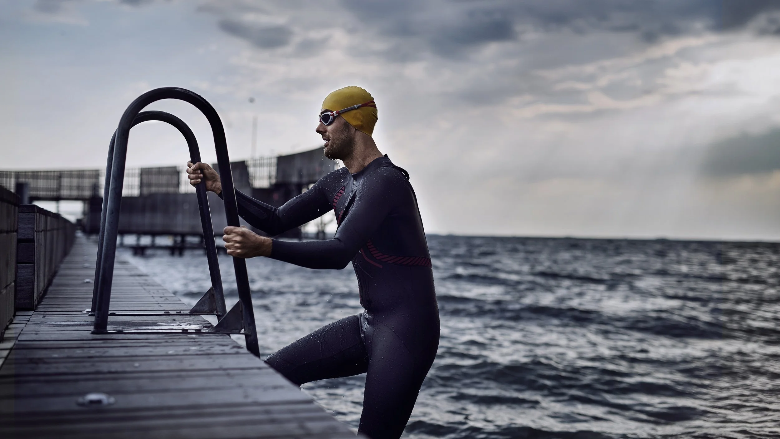 Male swimmer in black wetsuit, yellow swim cap, and goggles, holding onto a metal ladder on a wooden dock by the water, with dark cloudy sky in the background.
