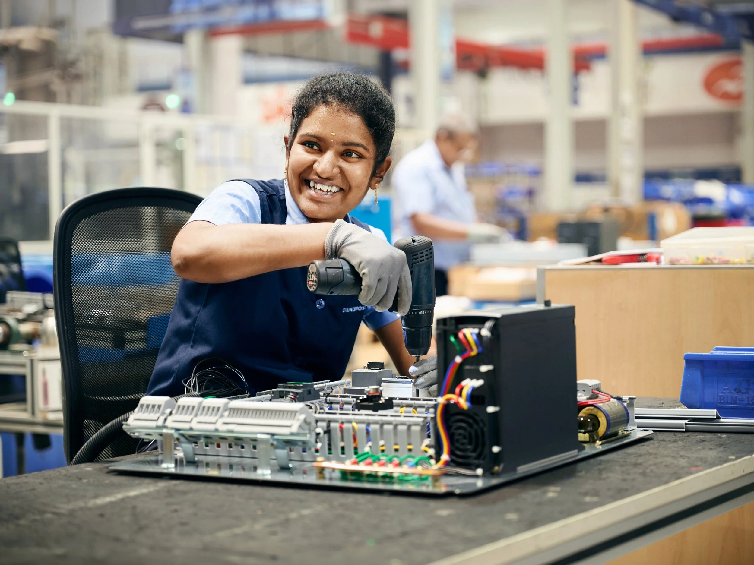 Worker assembling electronic components on production table, smiling during task
