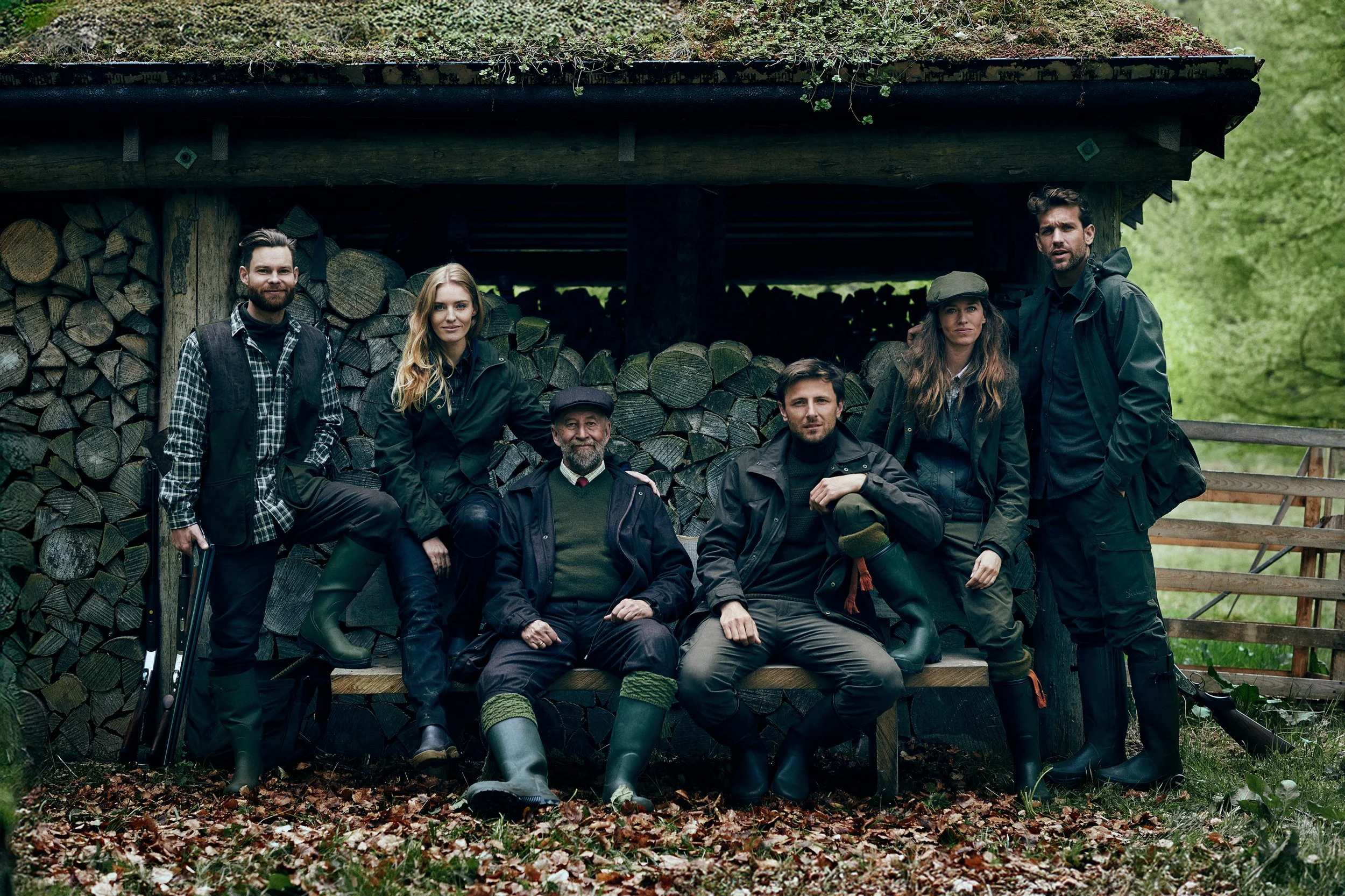 Group portrait of hunters across generations seated and standing by wood stack, traditional rural setting
