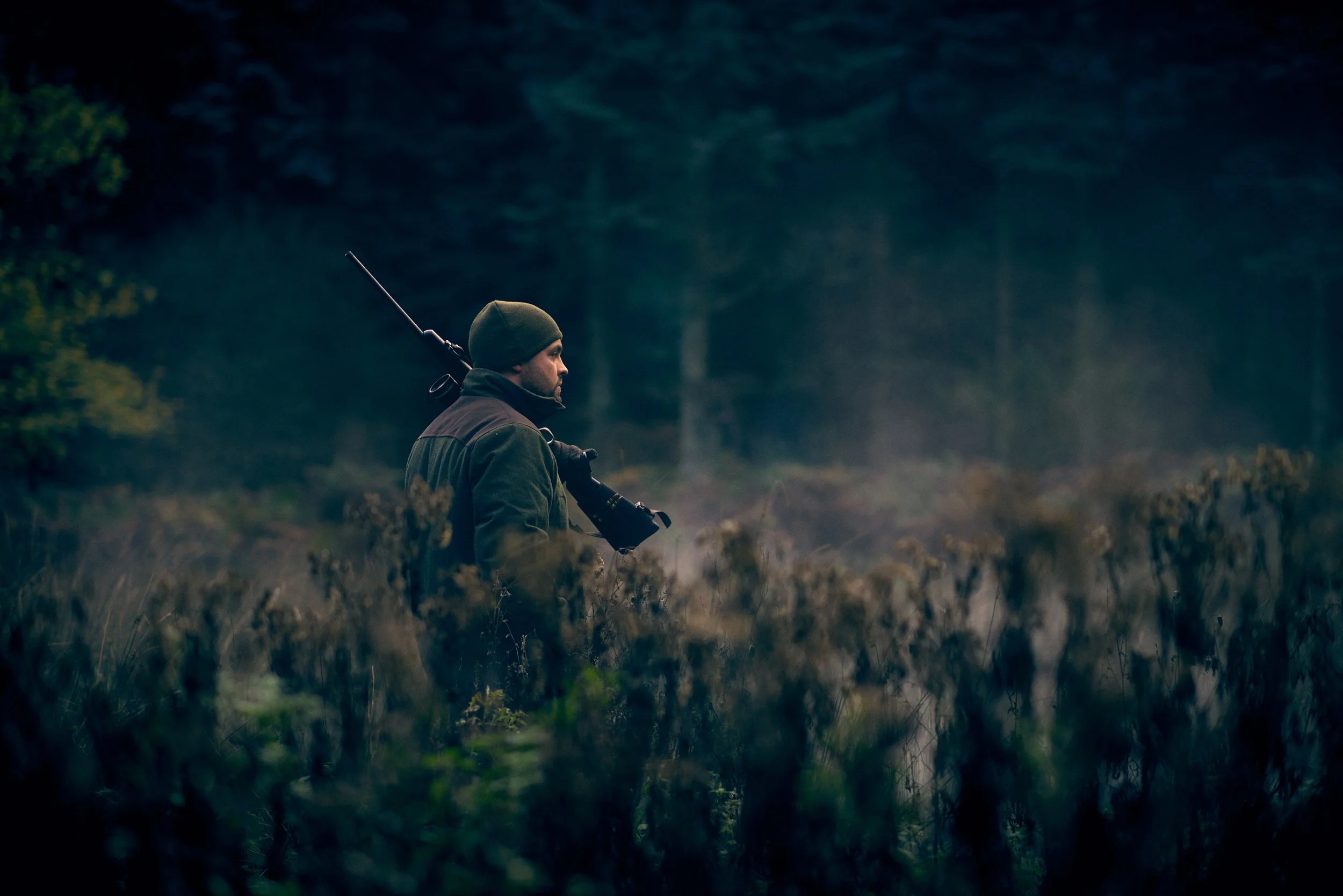 Hunter walking through tall vegetation at dusk, rifle over shoulder in quiet field setting