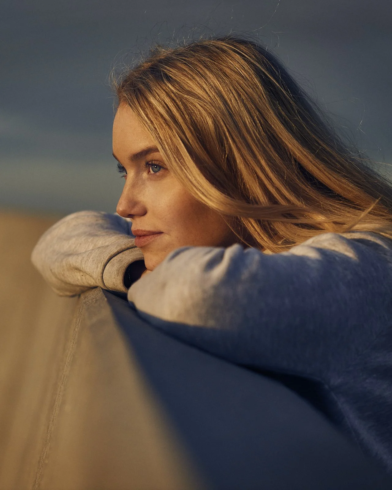 A young woman with long, light brown hair and blue eyes resting her chin on her arms, gazing thoughtfully into the distance against a blurred background.