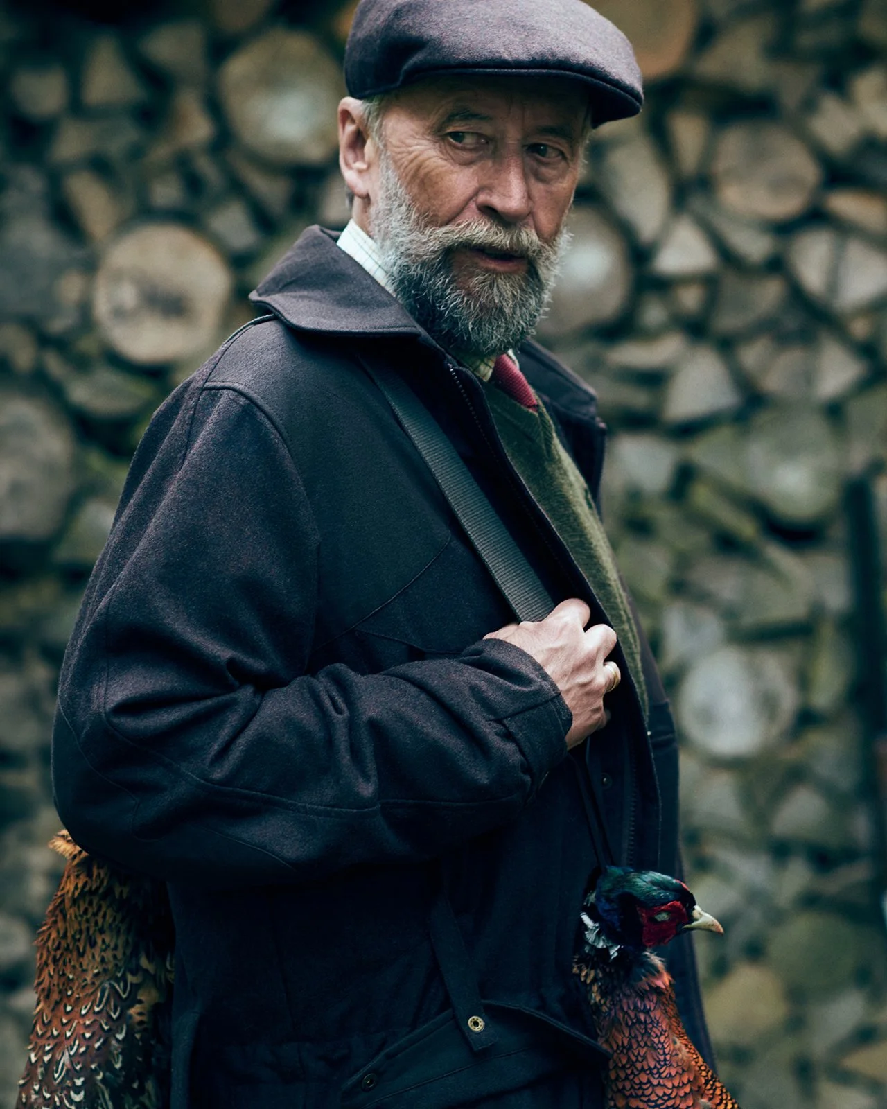An elderly man with a beard wearing a flat cap and dark jacket, holding a turkey, standing outdoors with stacked logs in the background.