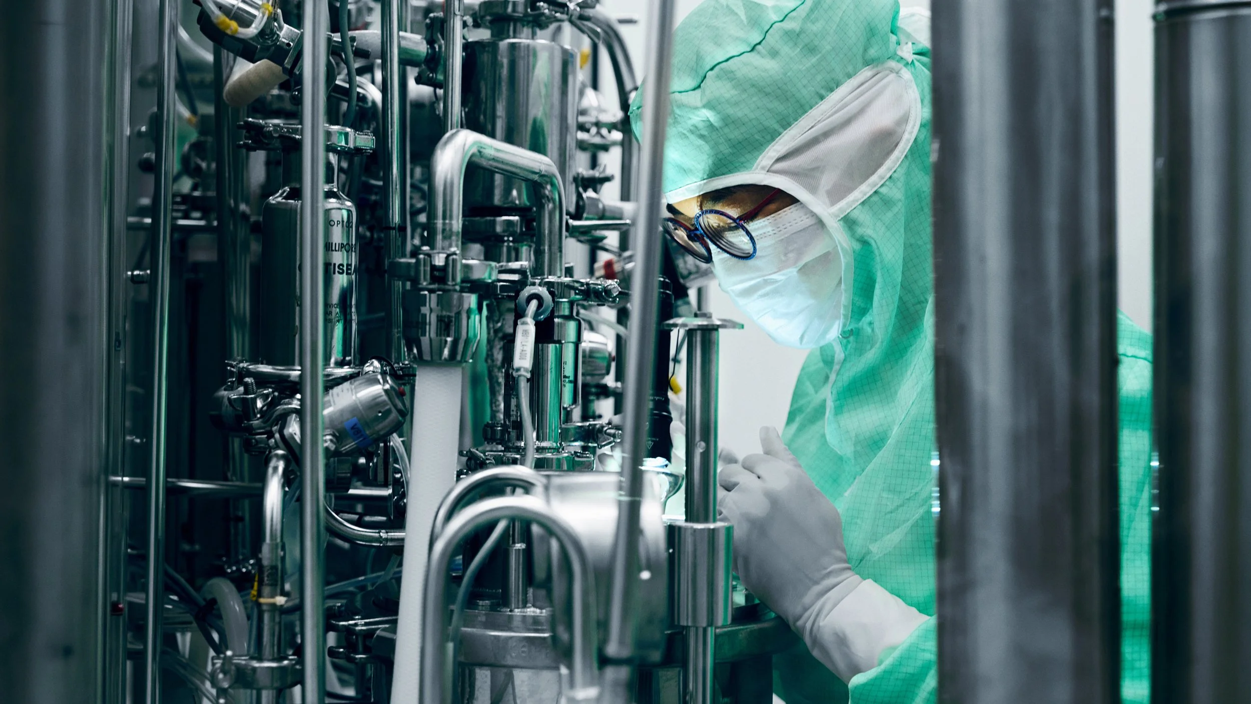 A scientist in protective gear working with complex stainless steel laboratory equipment in a cleanroom environment.