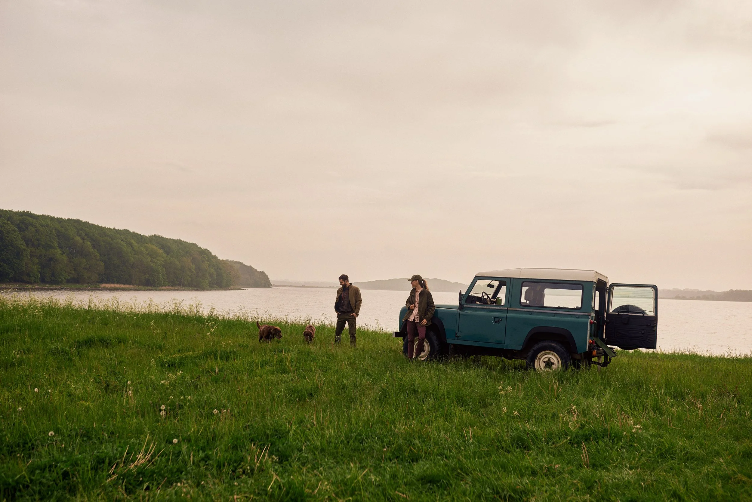 Two people standing by vehicle near lake with dogs, quiet moment in open landscape