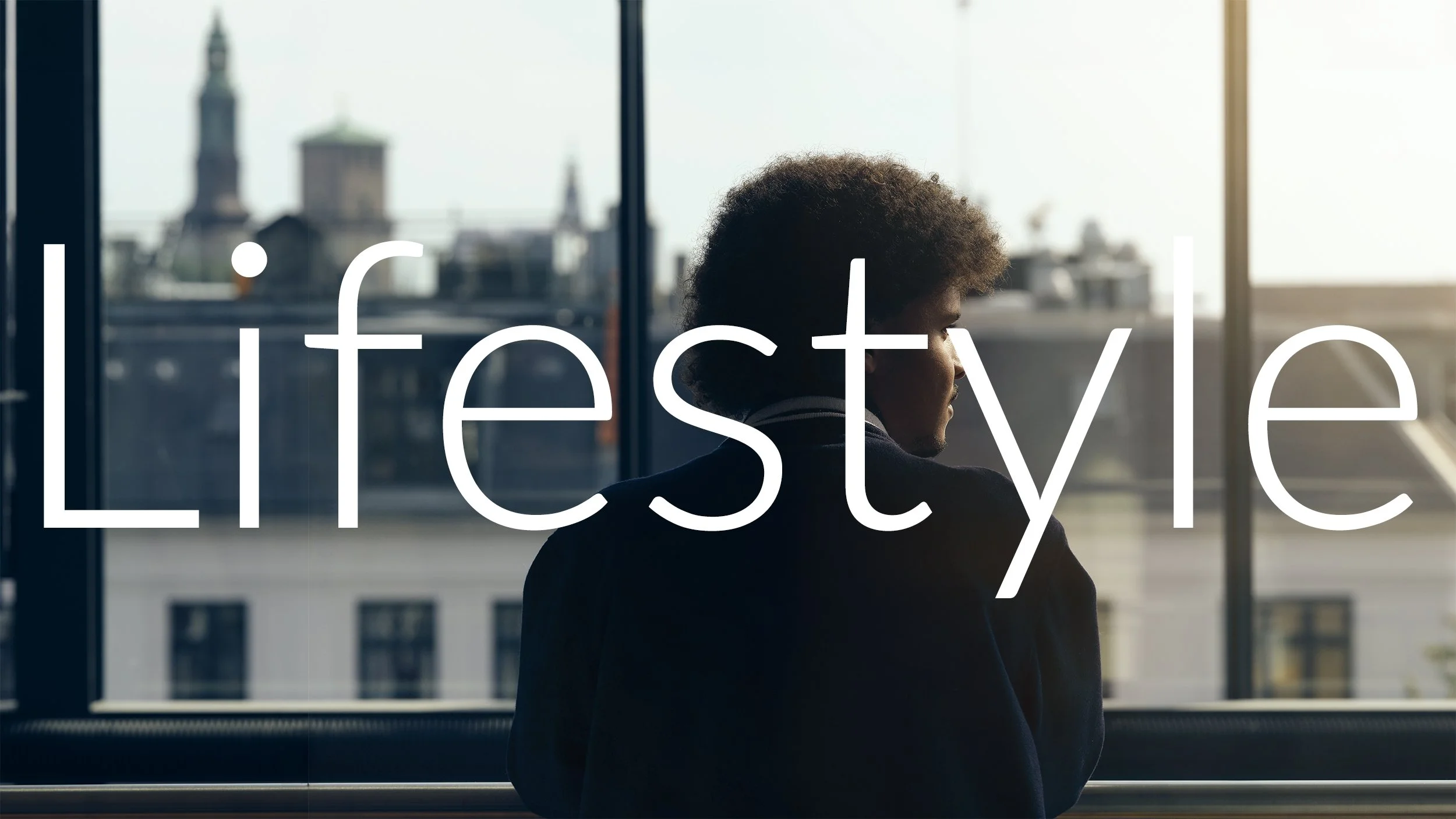 A person with curly hair looking out a window with a cityscape in the background. The word 'lifestyle' is written across the image.