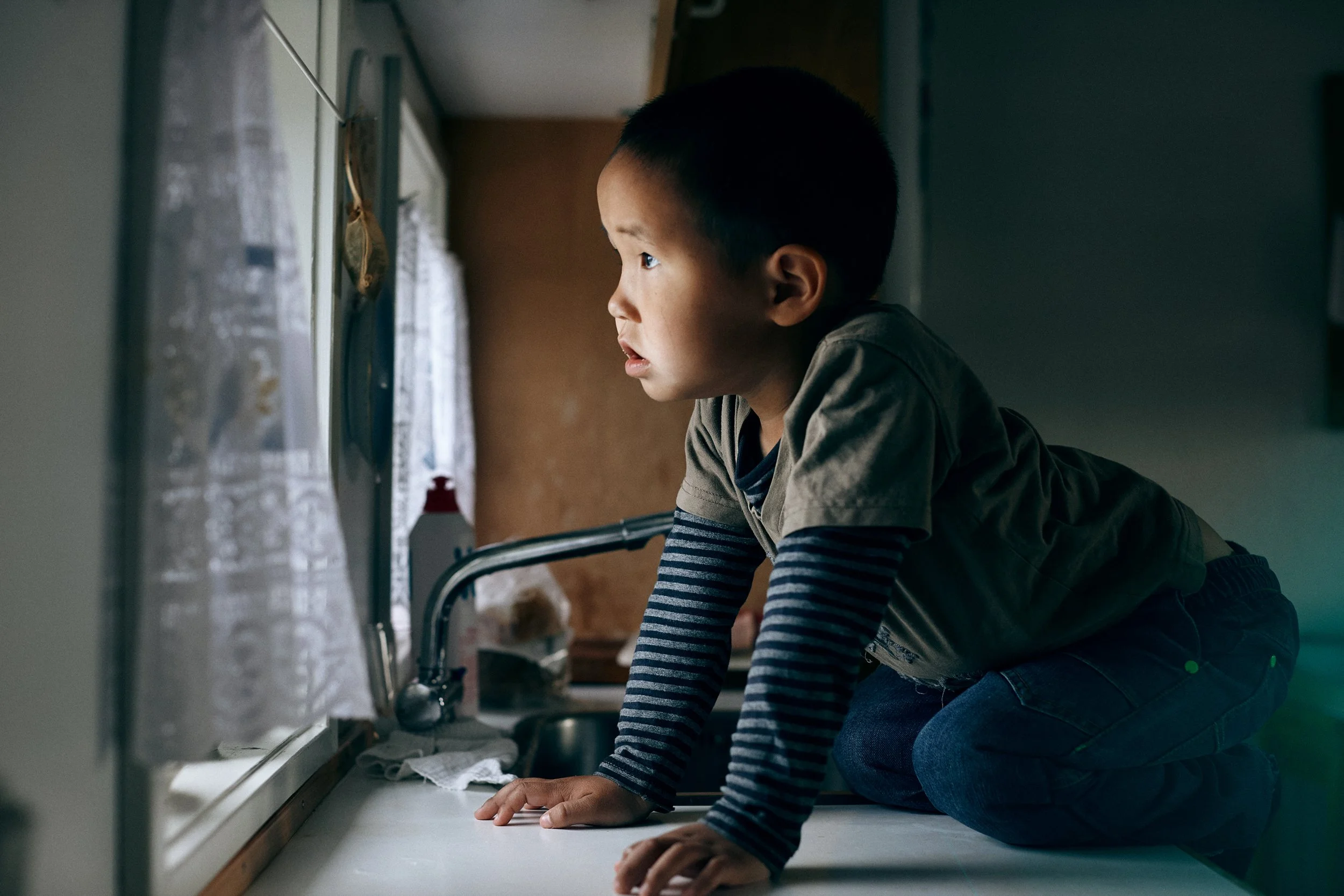 Child kneeling by window looking outside, soft light in domestic setting