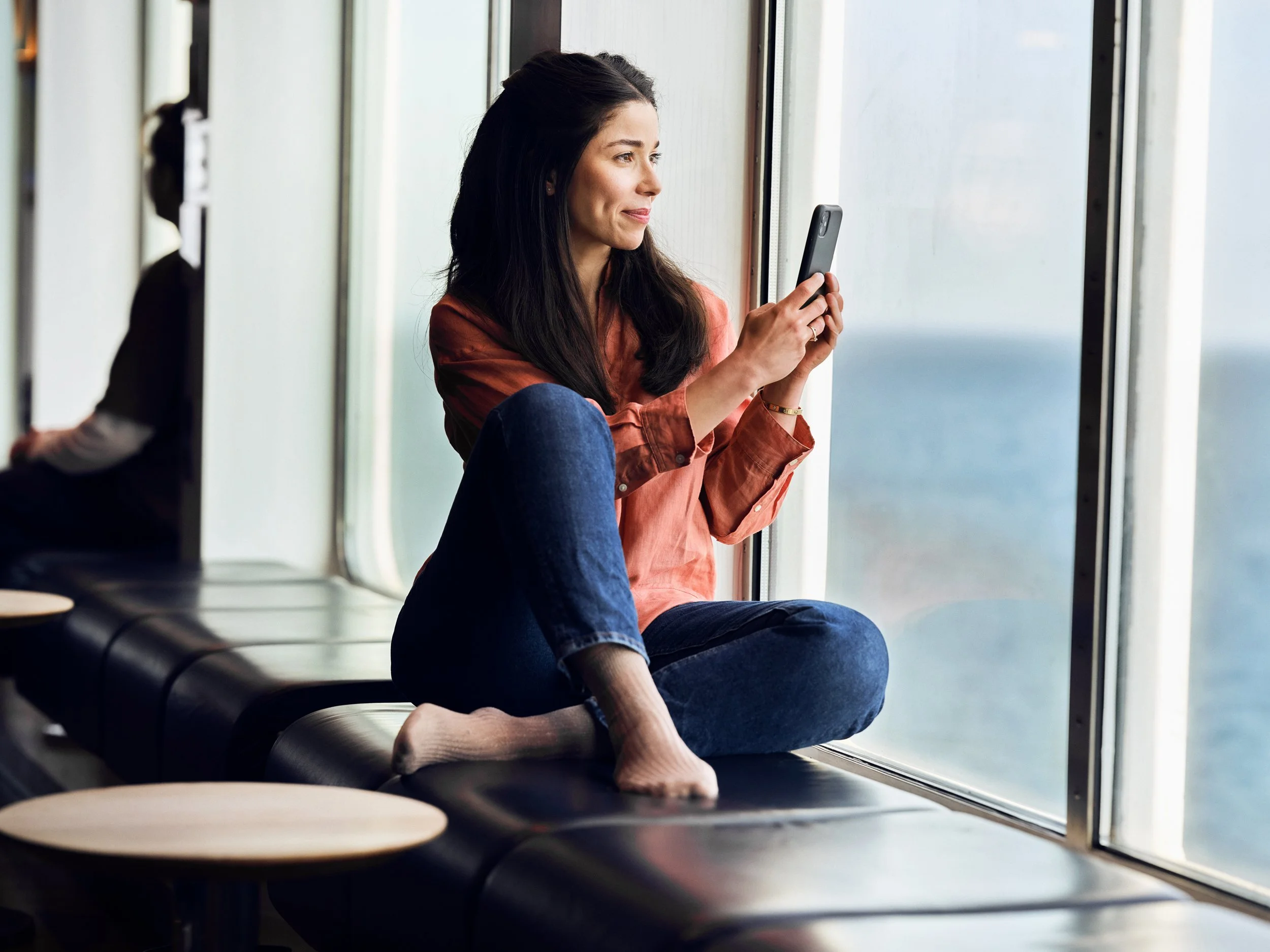 Woman sitting by window looking at phone, quiet moment overlooking sea