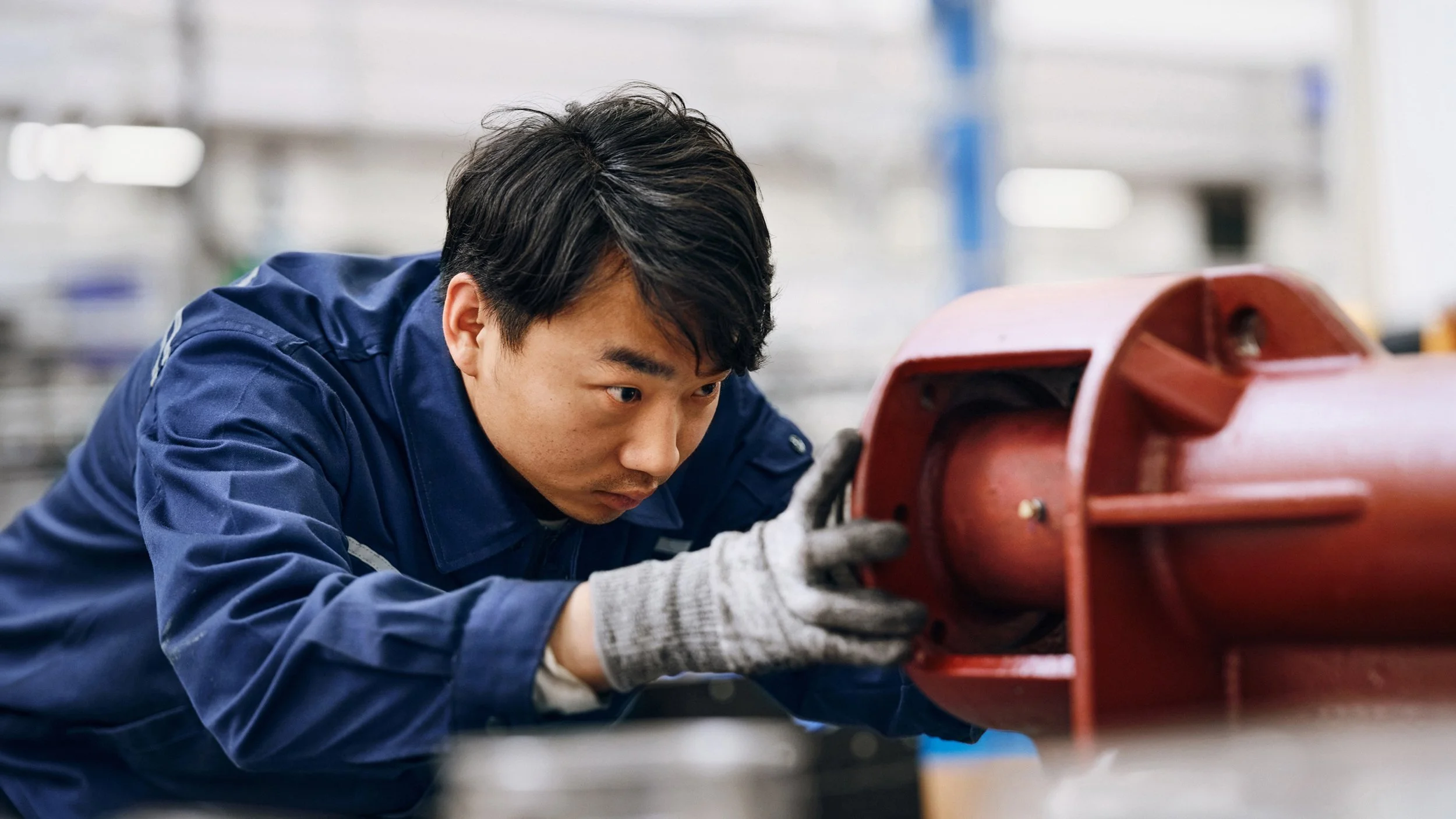 A worker in a blue uniform and gray gloves inspects or works on a red cylindrical object, possibly in a manufacturing or industrial setting.