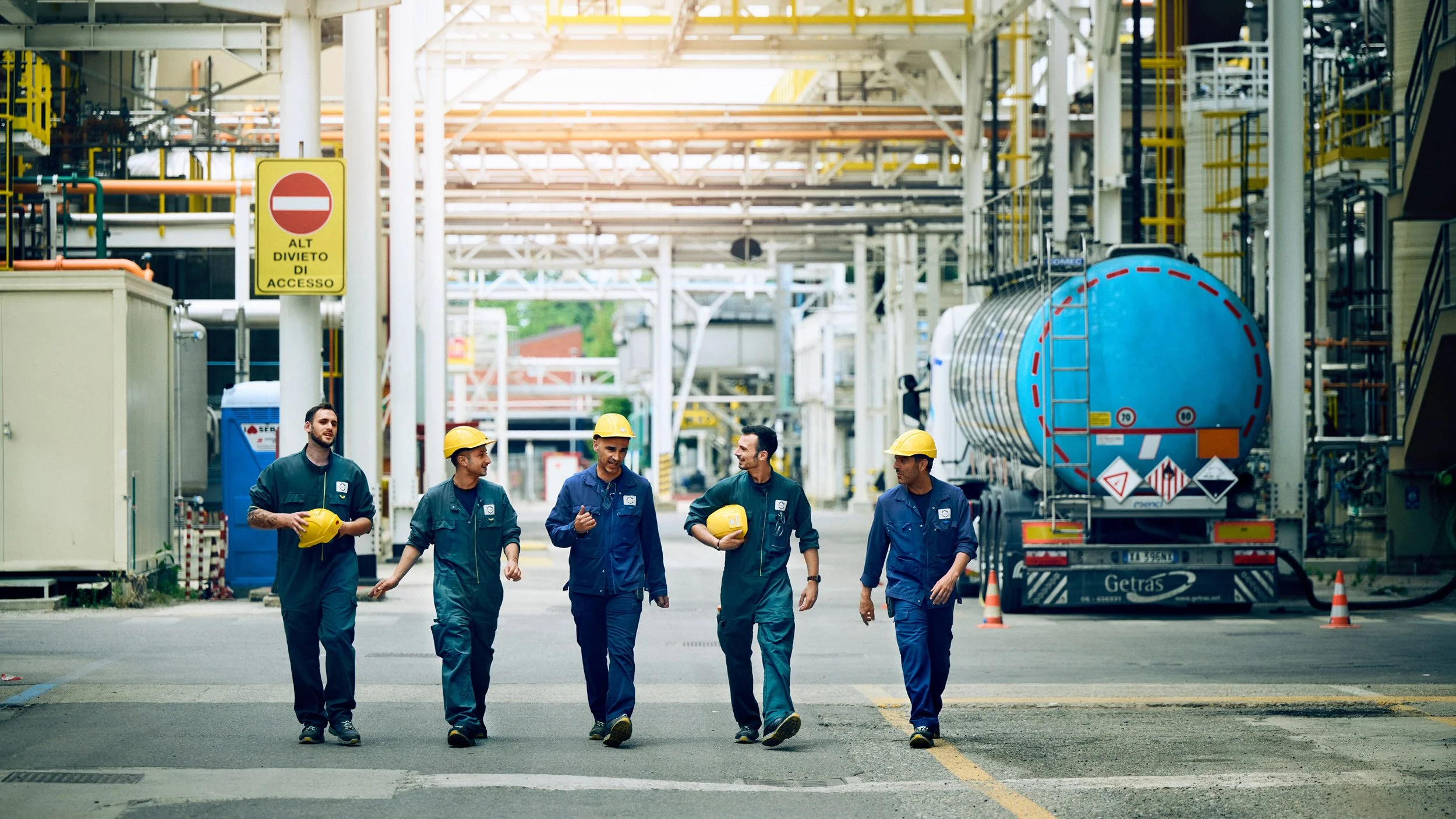 Five industrial workers, dressed in blue coveralls and yellow safety helmets, walking and conversing inside a factory or industrial site. One is holding a helmet, and they appear to be discussing something. The background features large pipes, a blue