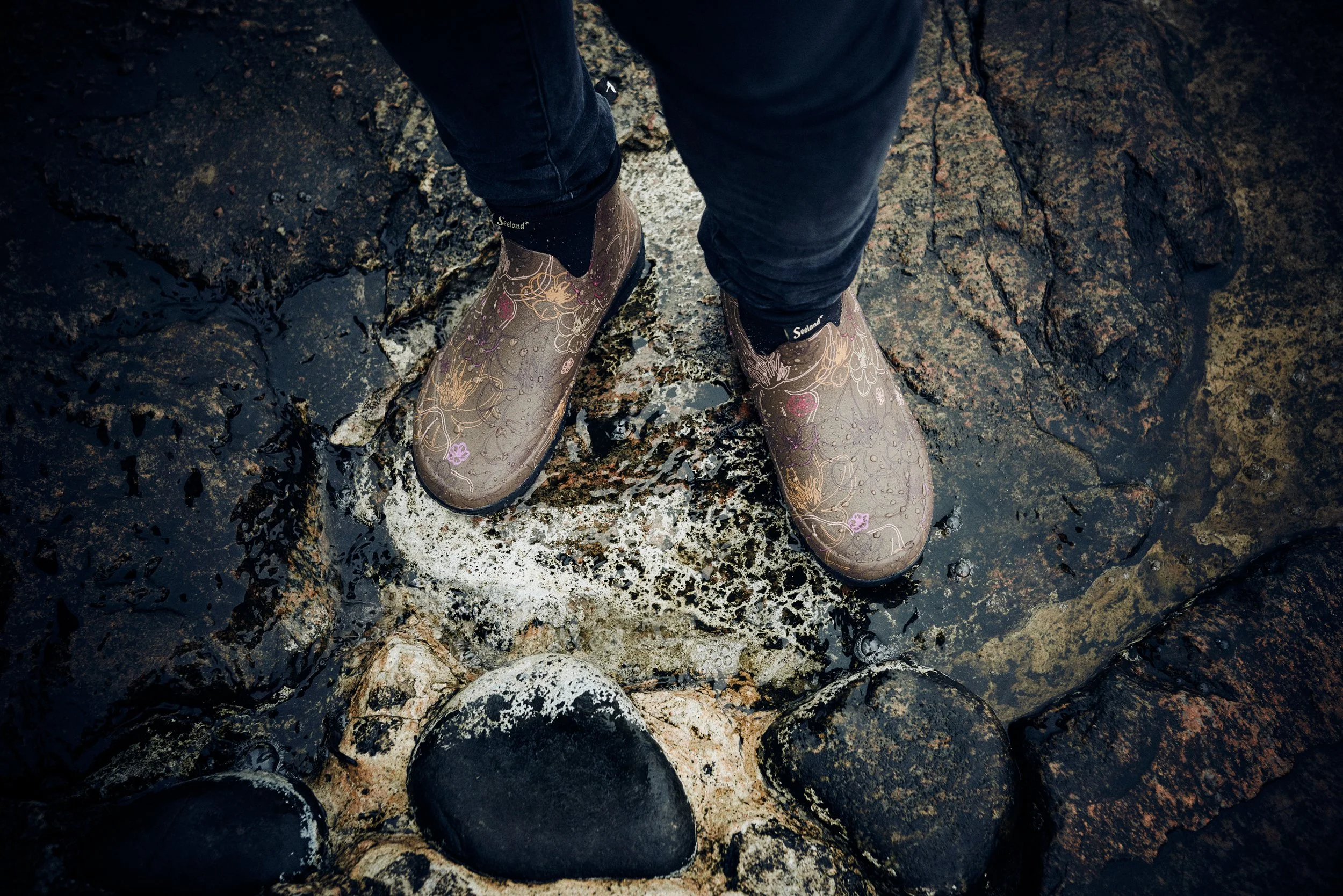 Close-up of boots standing on wet rocks by water, detail of coastal terrain
