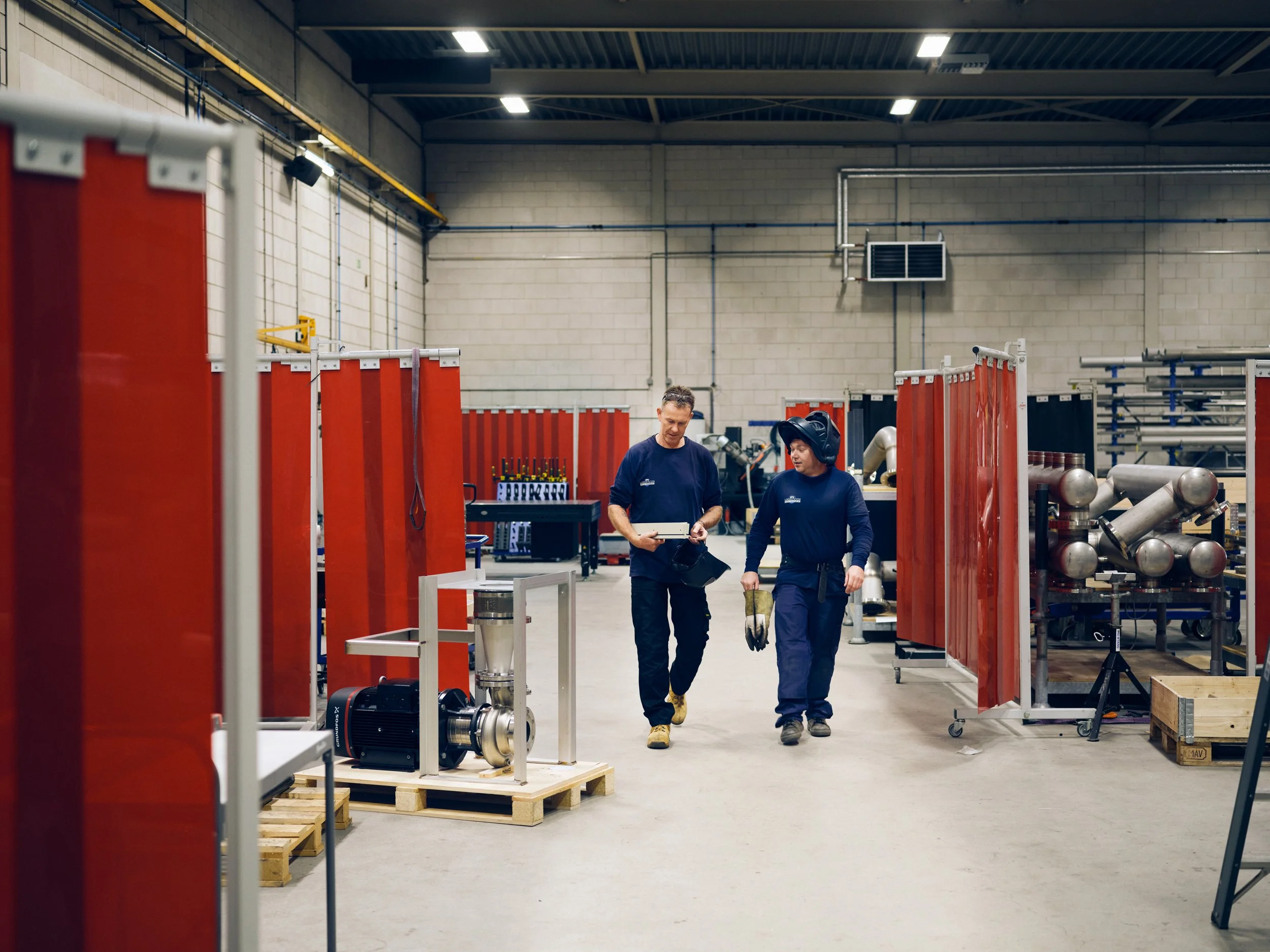 Two workers walking through fabrication workshop carrying equipment
